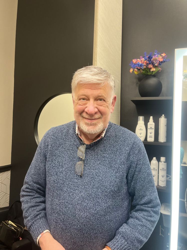 A smiling person in a blue textured sweater stands in a salon with hair products on shelves in the background.