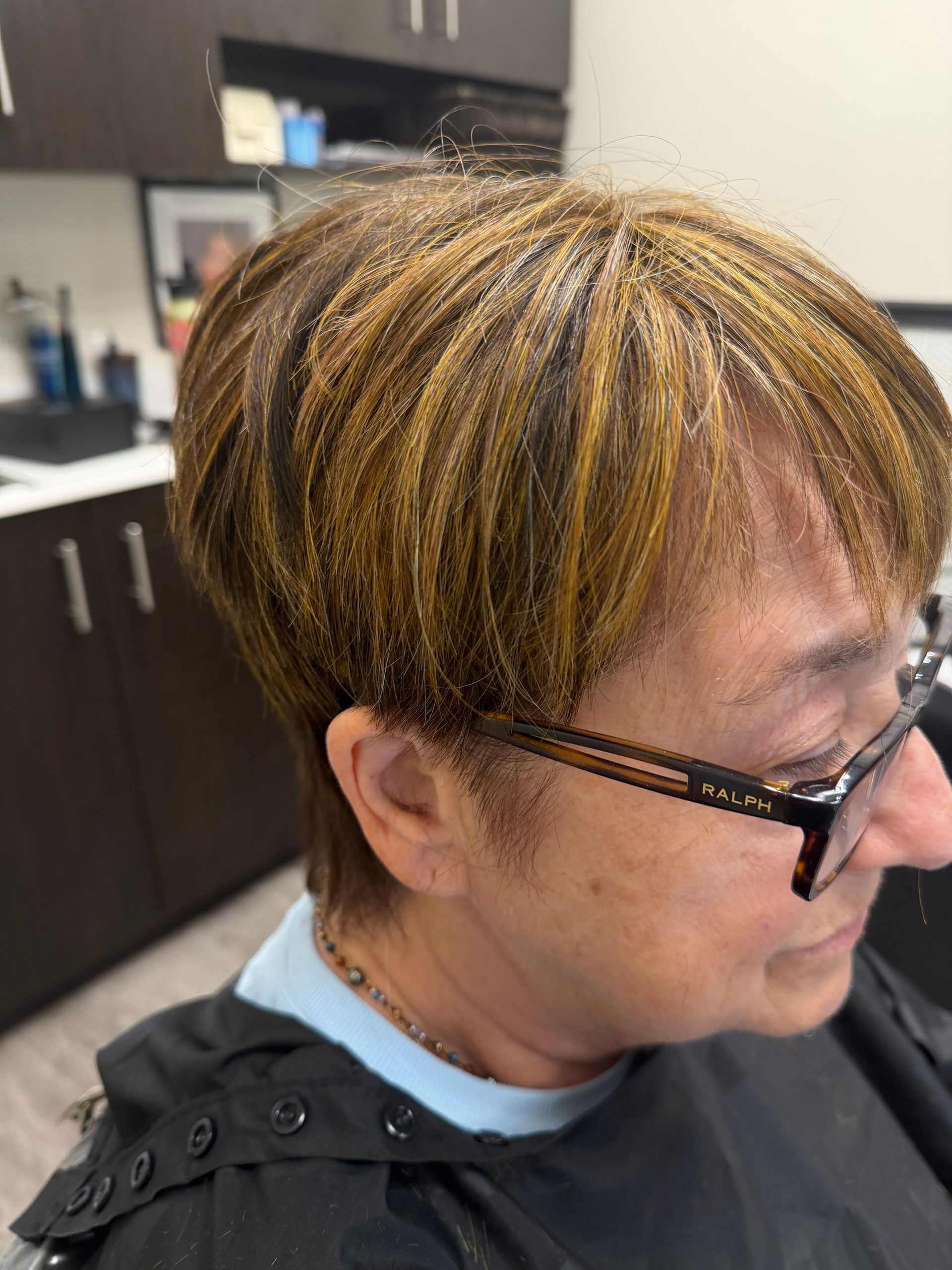 Person with short highlighted hair and glasses in a salon chair, side profile indoors
