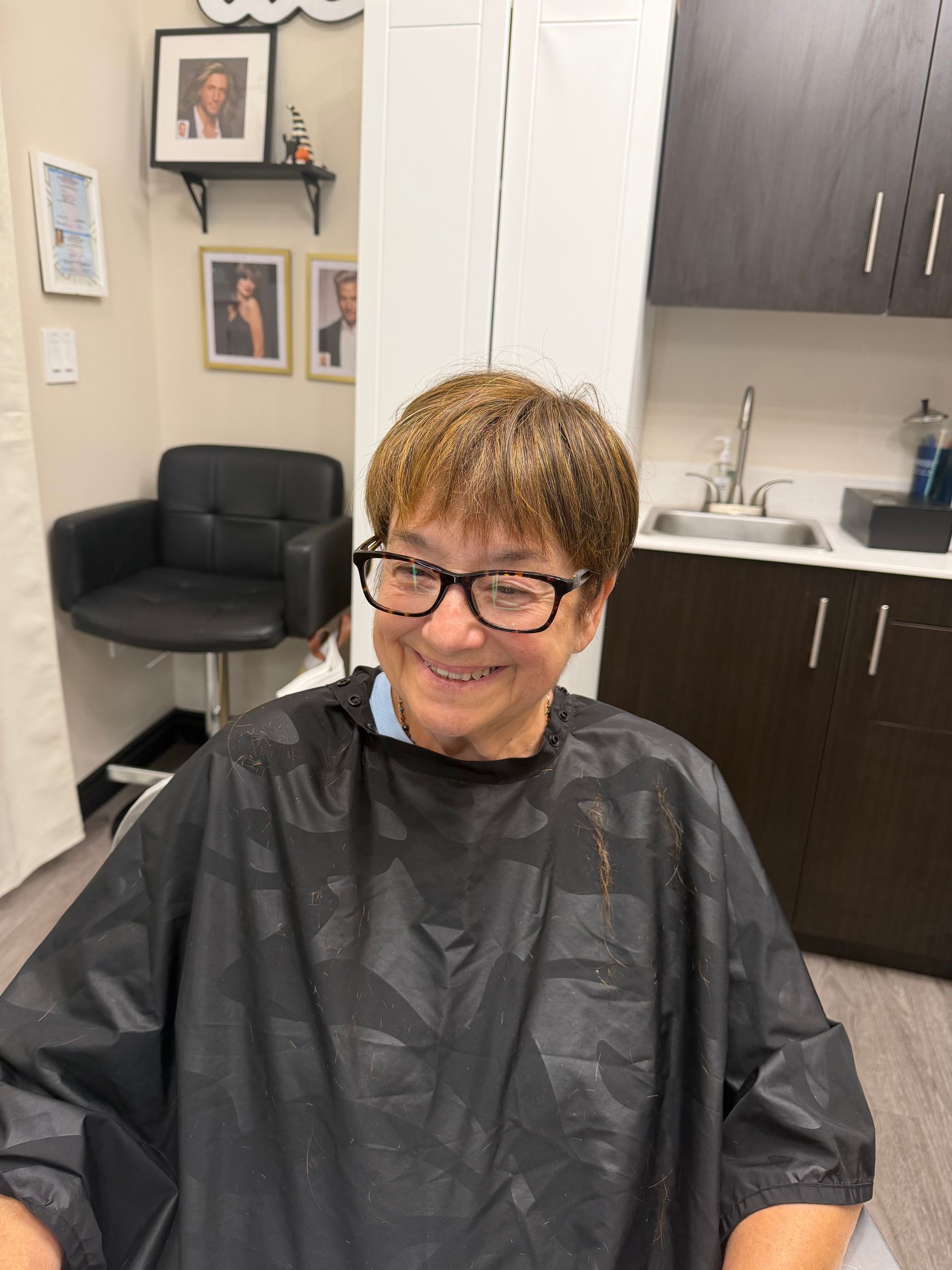 Person smiling in a salon chair wearing a black cape, with framed photos and dark cabinets in the background.