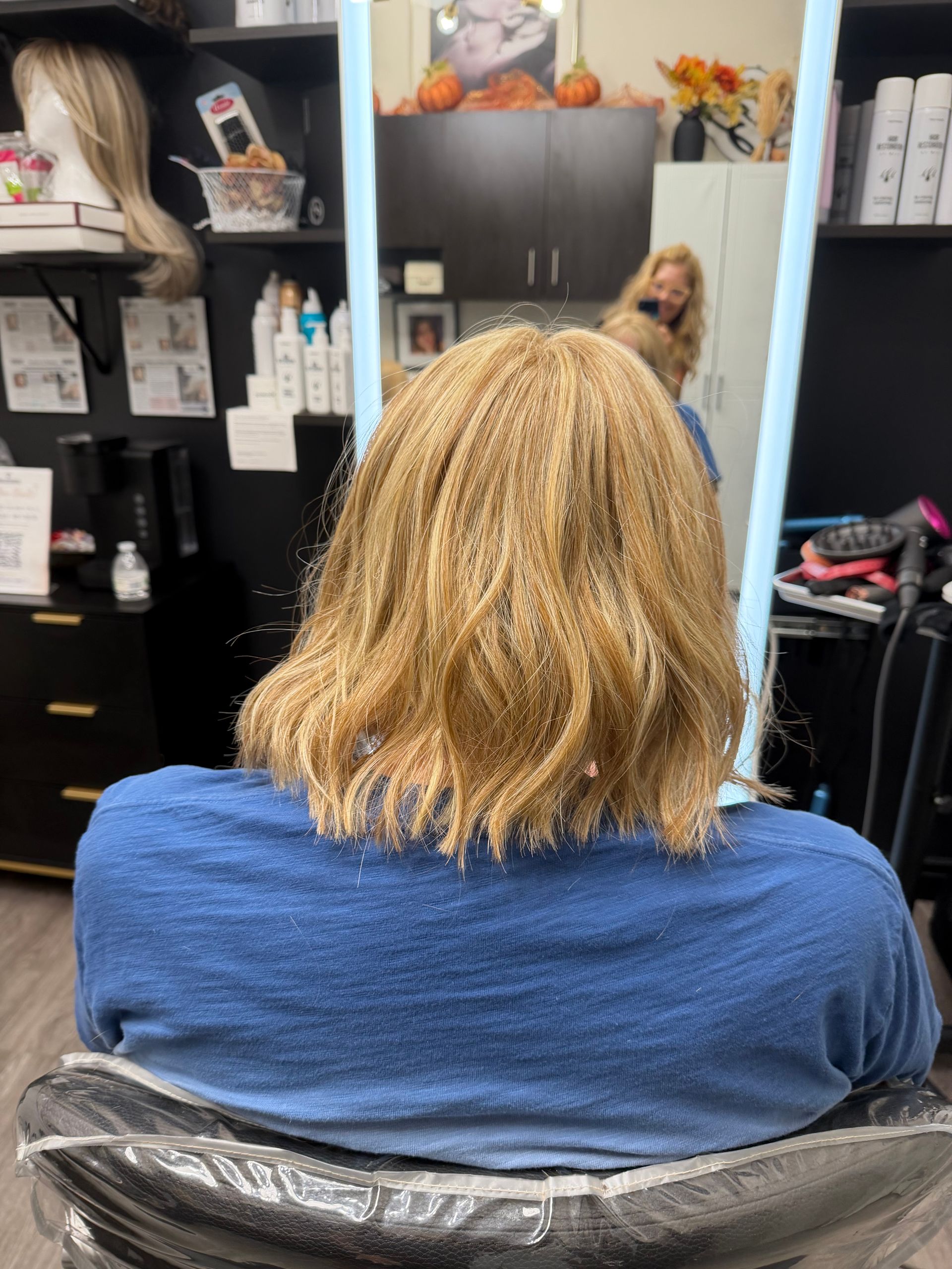 A person with a mustache smiles while sitting in a chair inside a hair salon with shelves of products in the background.