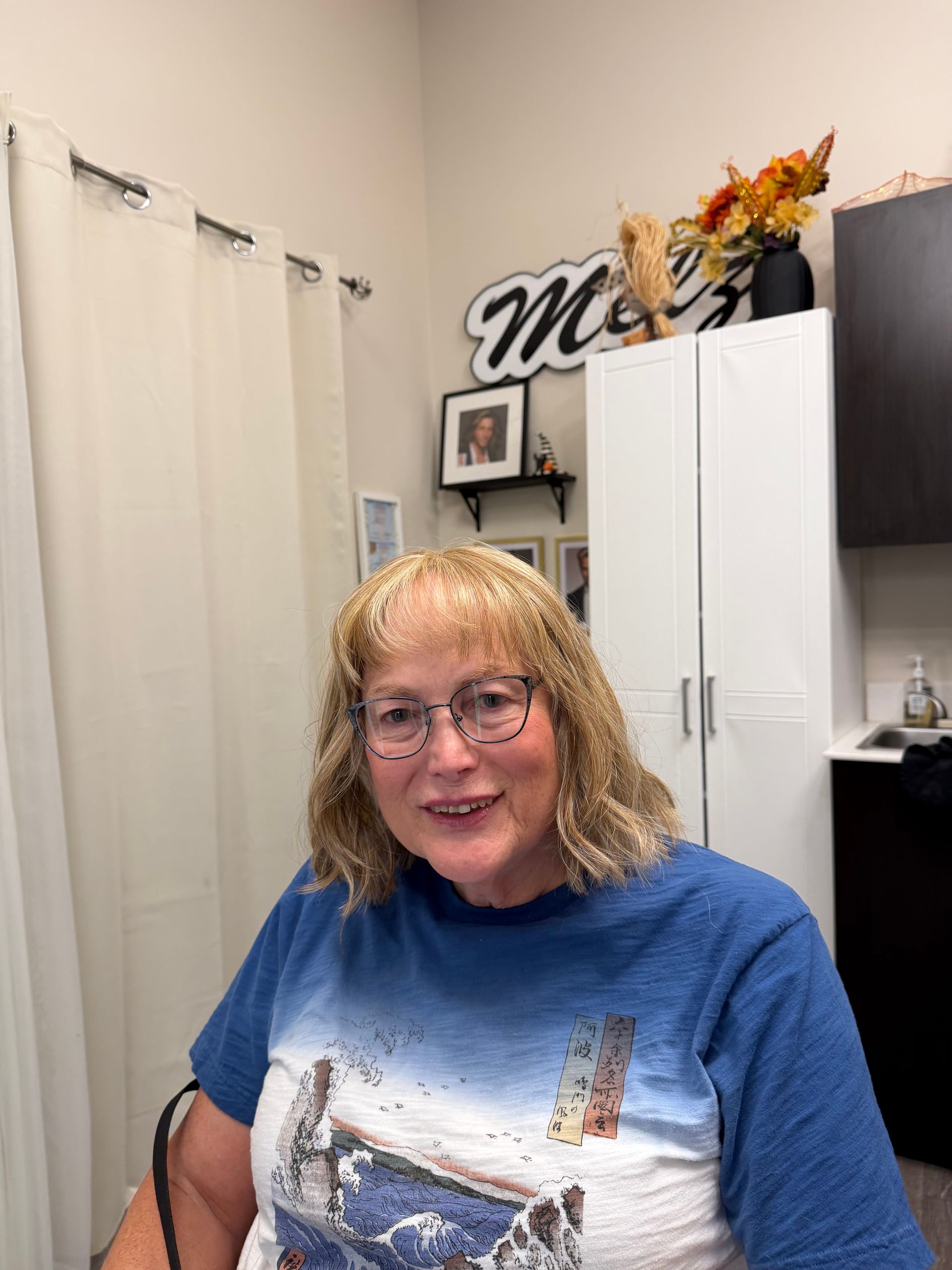 Woman smiling in a home office, wearing glasses and a blue graphic shirt, with white cabinets behind her.