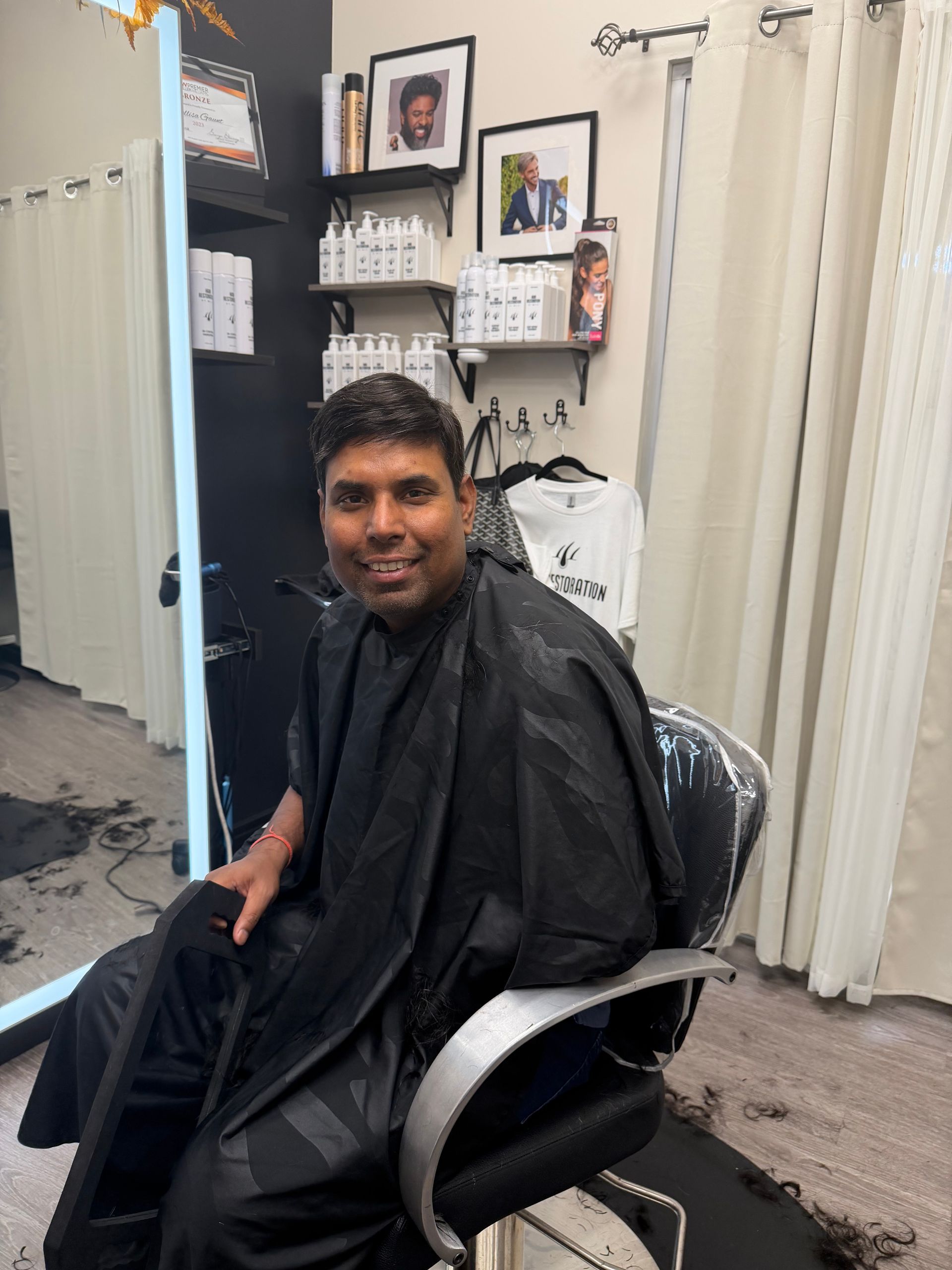 Man sitting in a salon chair wearing a black cape, smiling in front of a mirror and shelves with hair products.