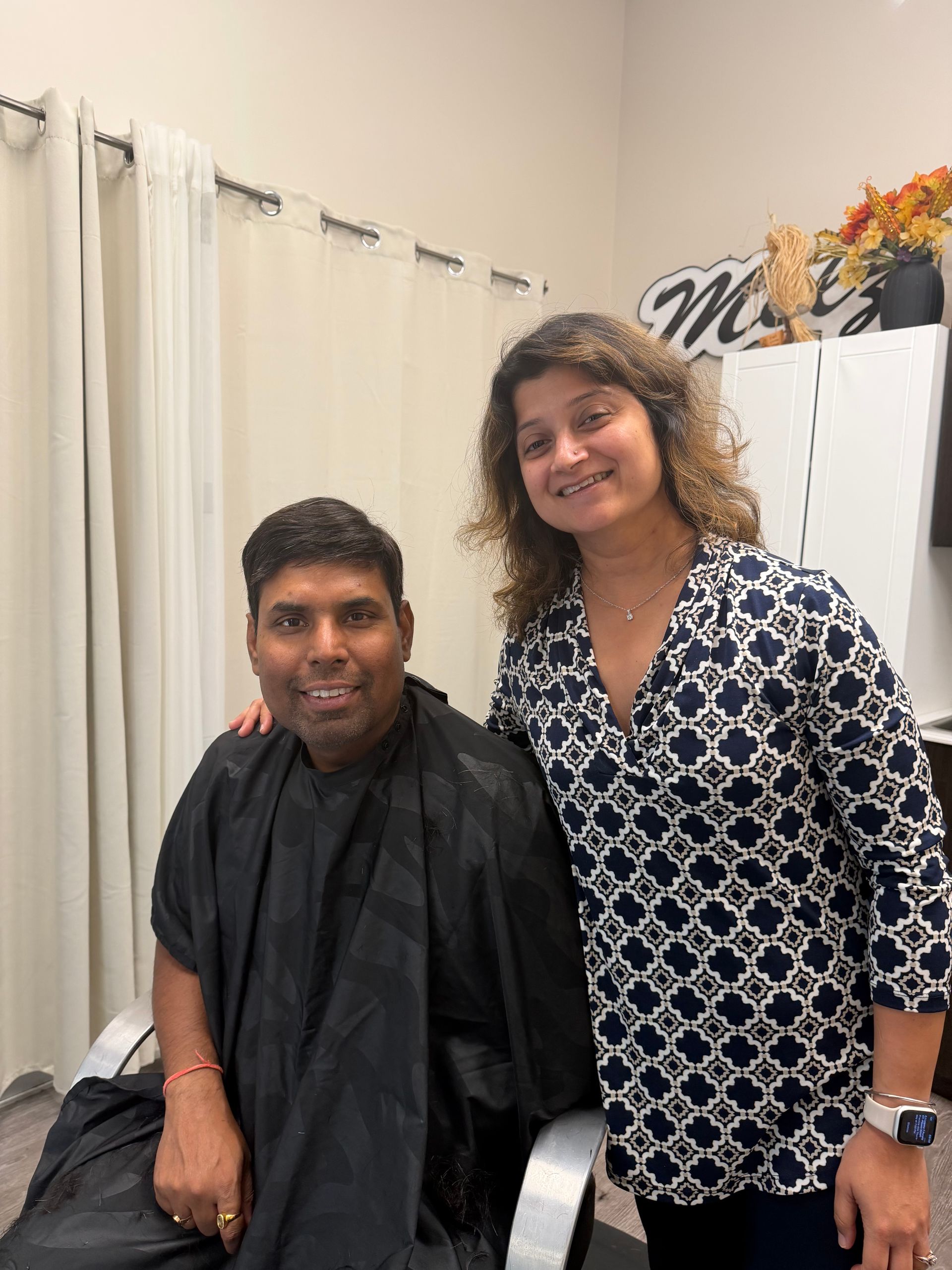 Two people posing in a salon, one seated in a black cape and one standing beside them smiling.