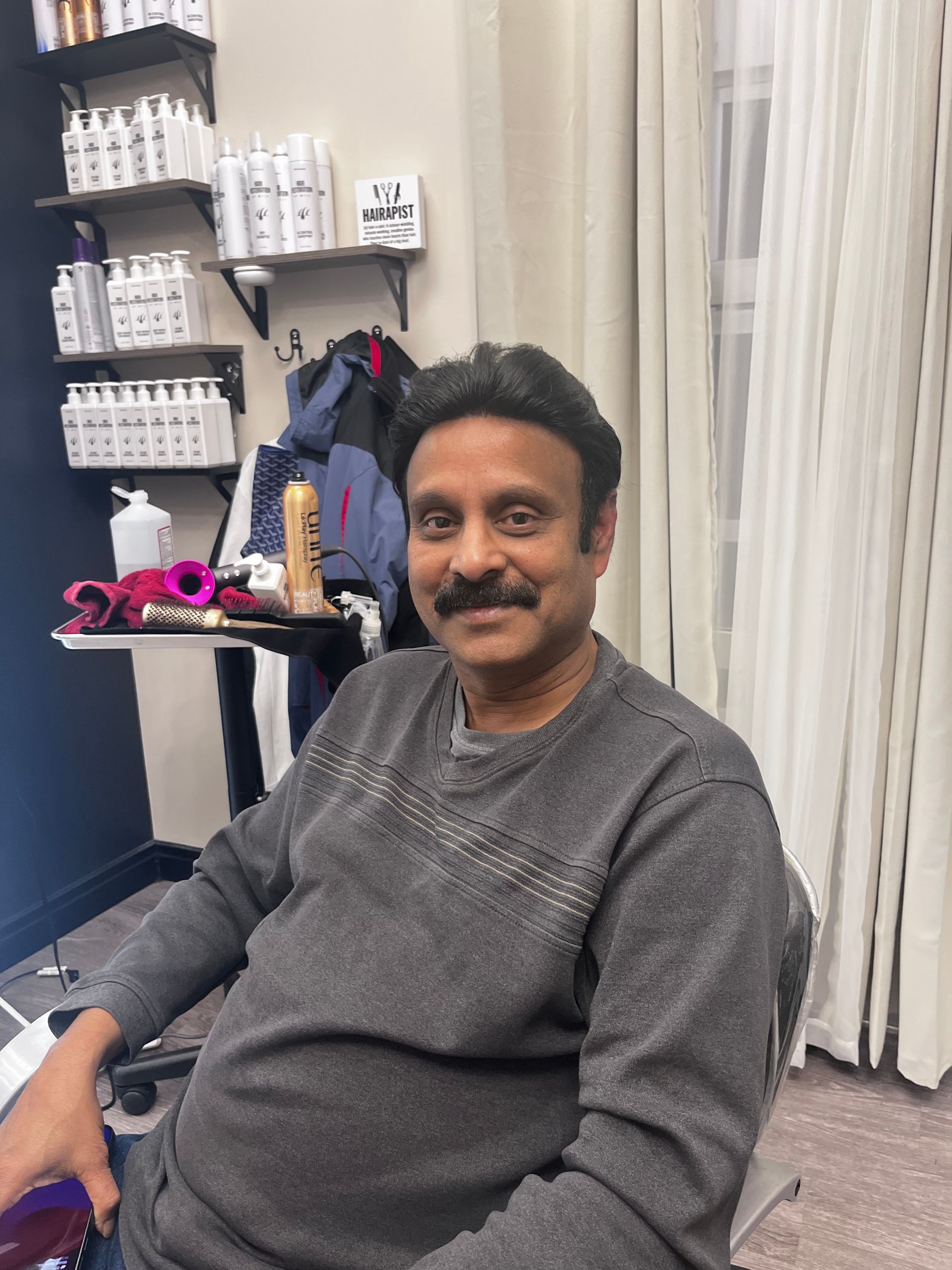 A person with a mustache smiles while sitting in a chair inside a hair salon with shelves of products in the background.
