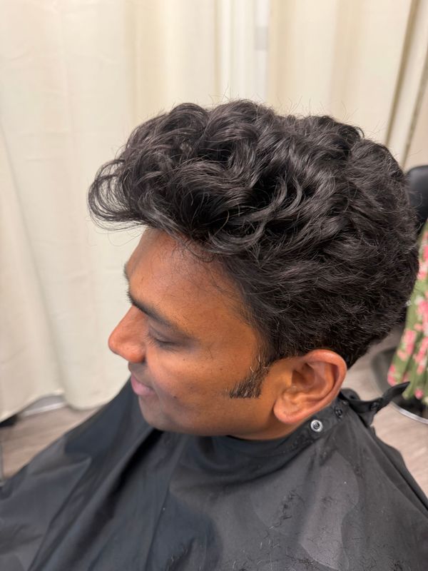 Side-profile of a person with curly hair wearing a black salon cape, seated indoors near curtains.