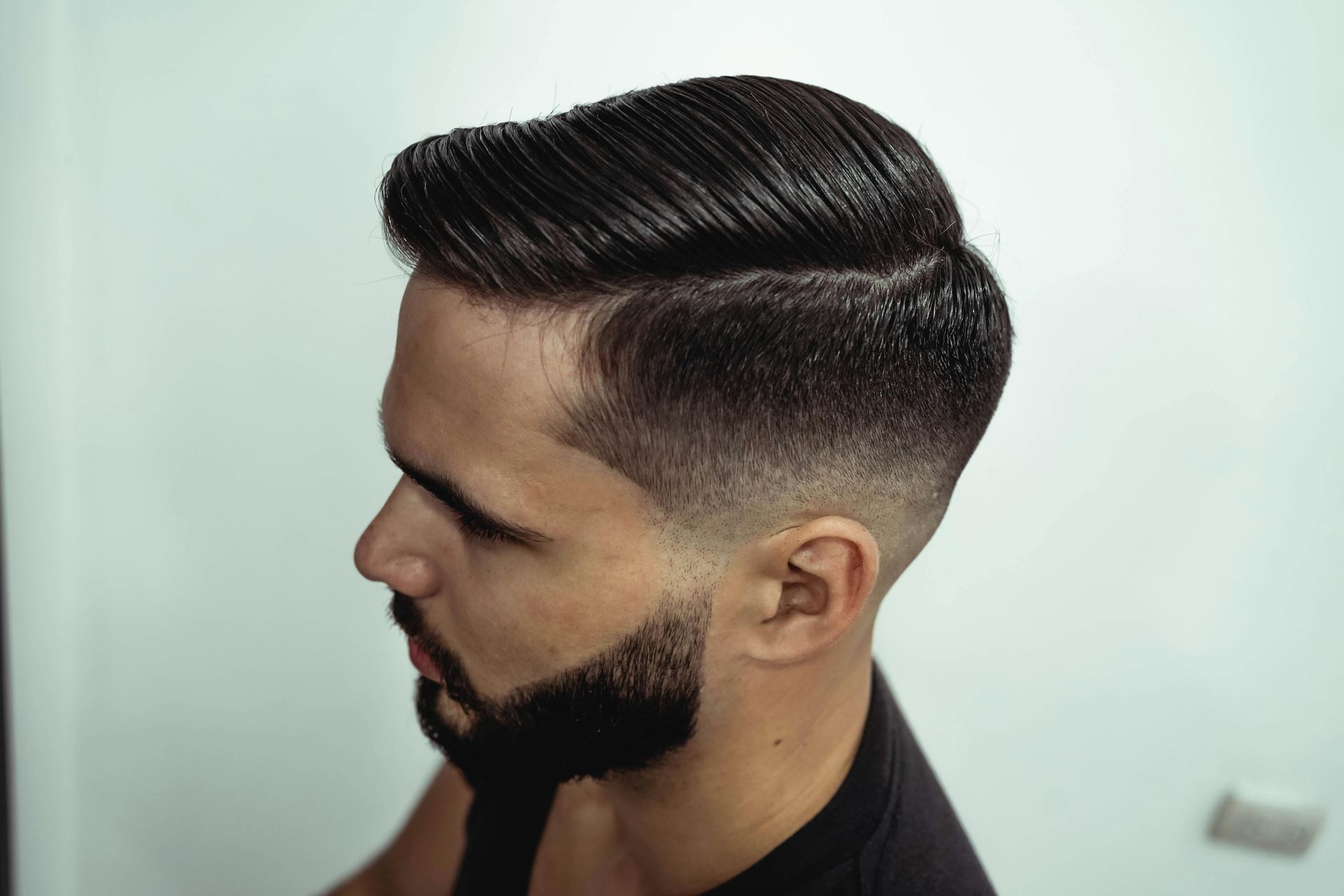 A close-up of a man with a side-parted hairstyle and a skin fade on the sides, paired with a short, neatly groomed beard.