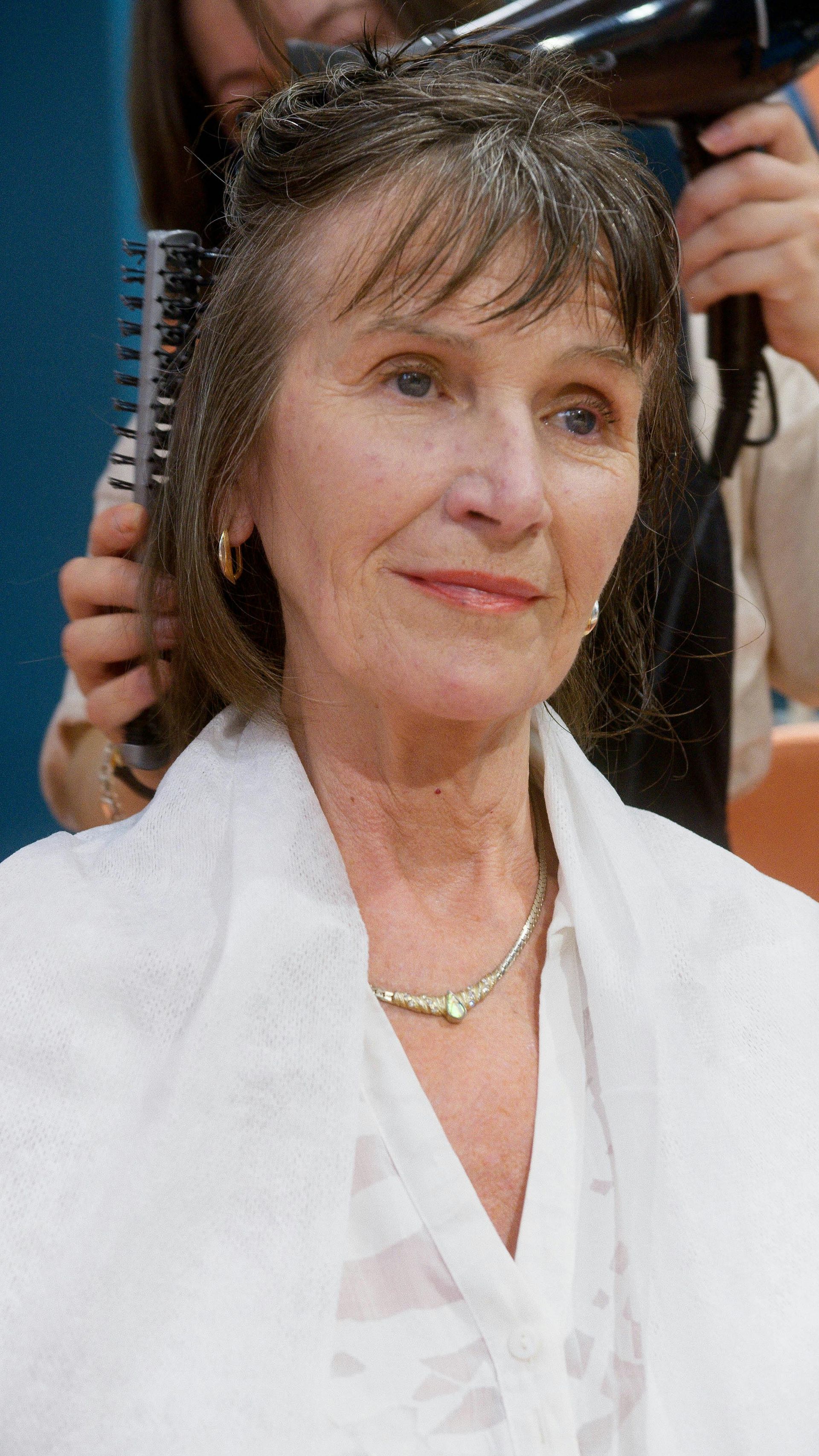 Woman getting hair styled, wearing a white shawl. Someone uses a brush and a hair dryer.