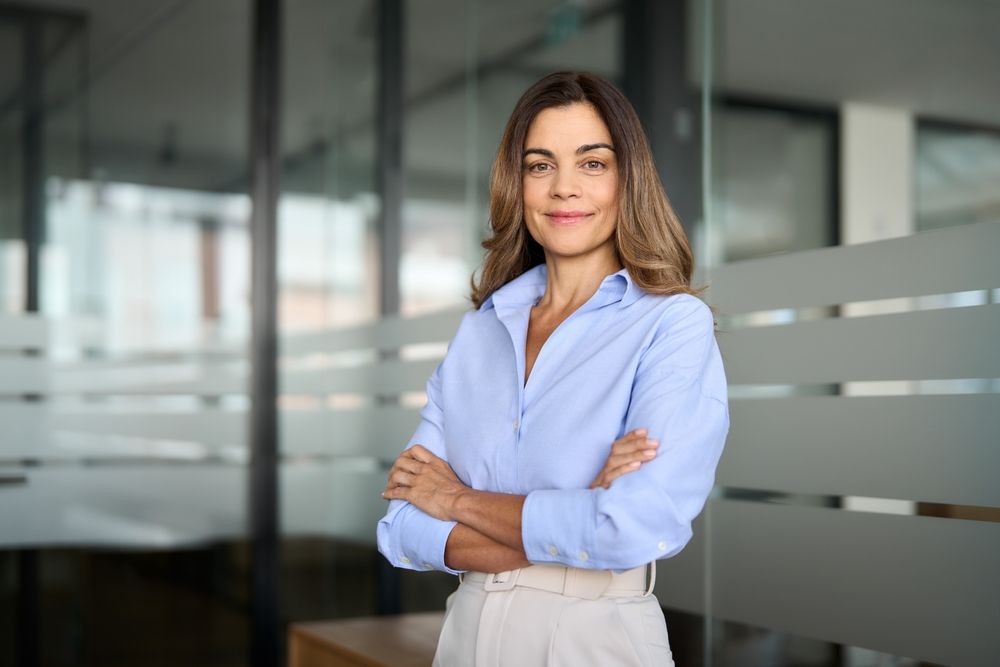Woman with arms crossed, wearing light blue shirt and tan pants, smiling in an office setting.