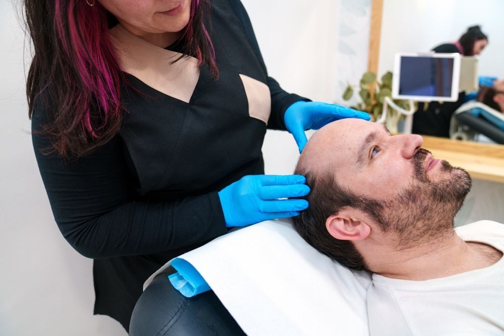 A woman in blue gloves examining a man's head, likely for hair treatment.