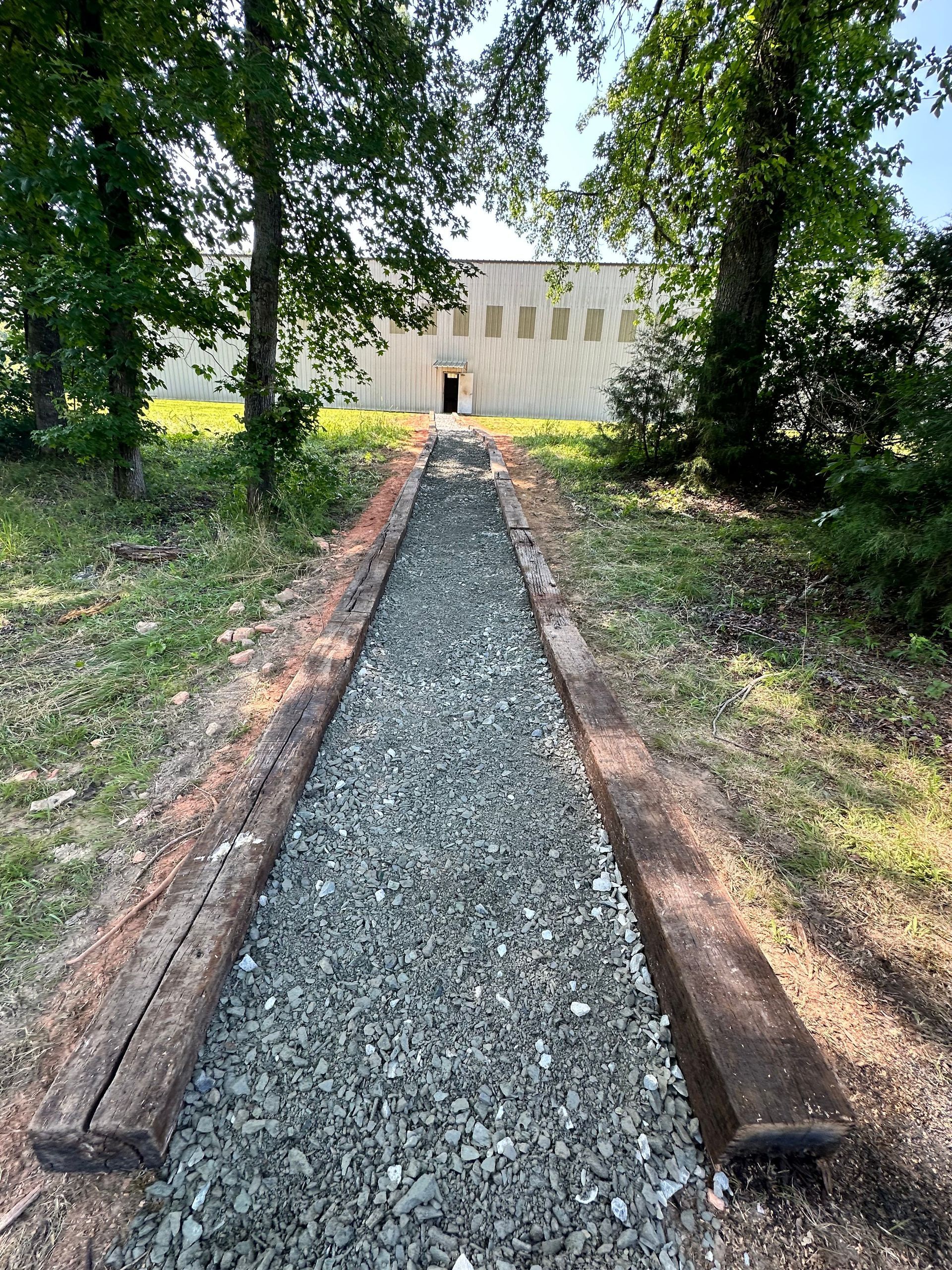 Pathway of gravel and wood leading to a light-colored building, trees on either side.