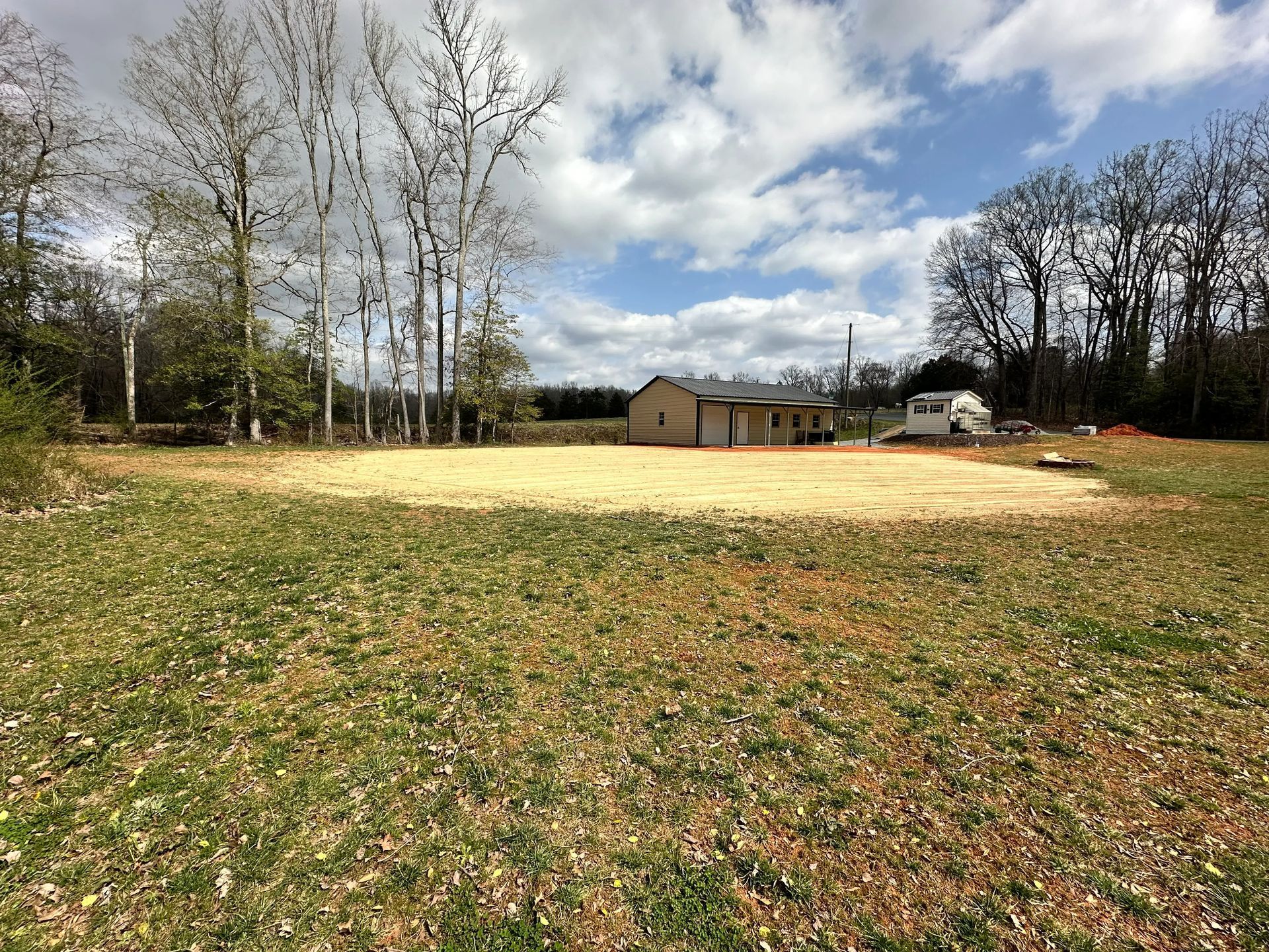 A clearing with a tan surface, surrounded by grass, trees, and a small building under a cloudy sky.