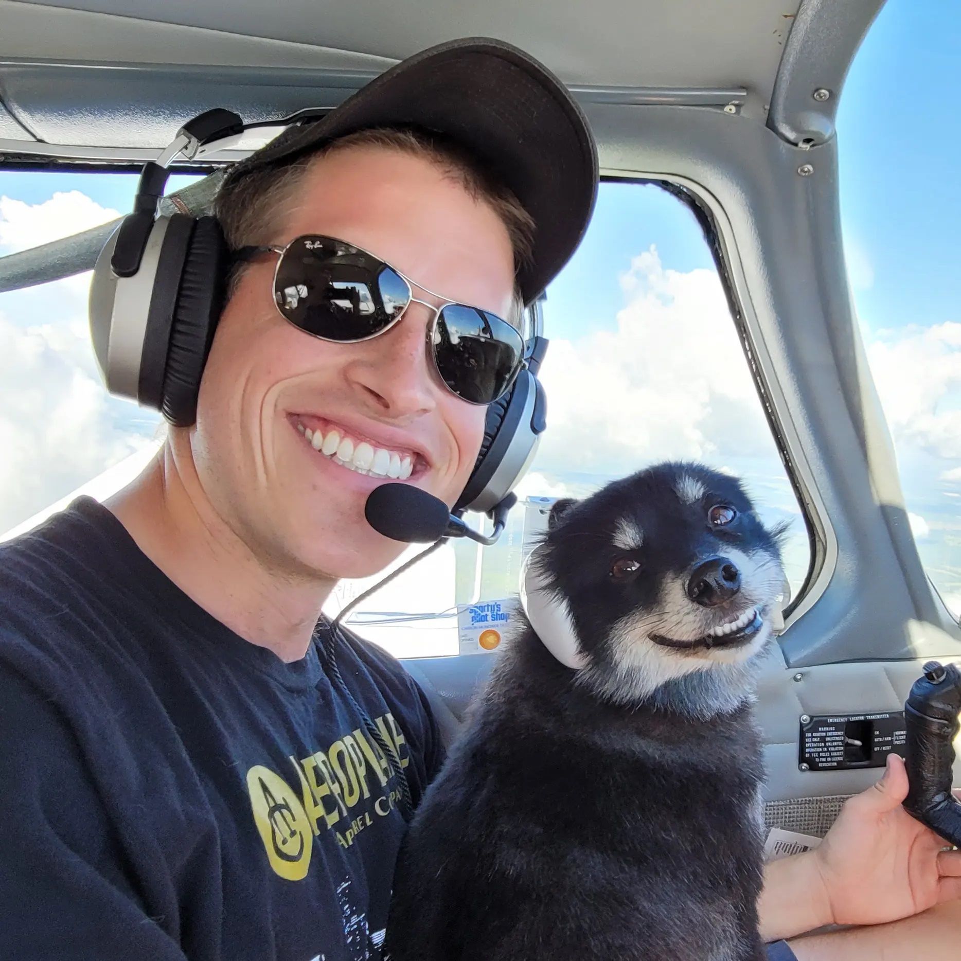 Man and dog in airplane cockpit, both wearing headsets, smiling. Blue sky visible.