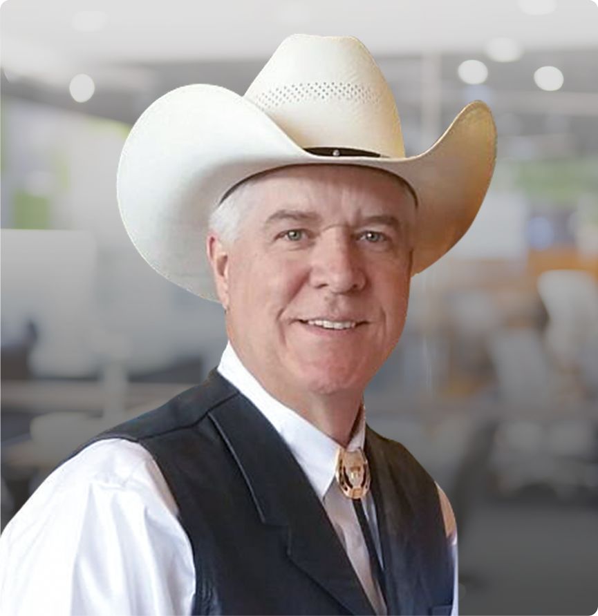 Man in cowboy hat and vest, bolo tie, smiling. Indoors, blurred background.