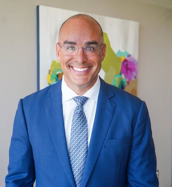 Smiling man in a blue suit and tie standing indoors in front of a colorful wall art piece