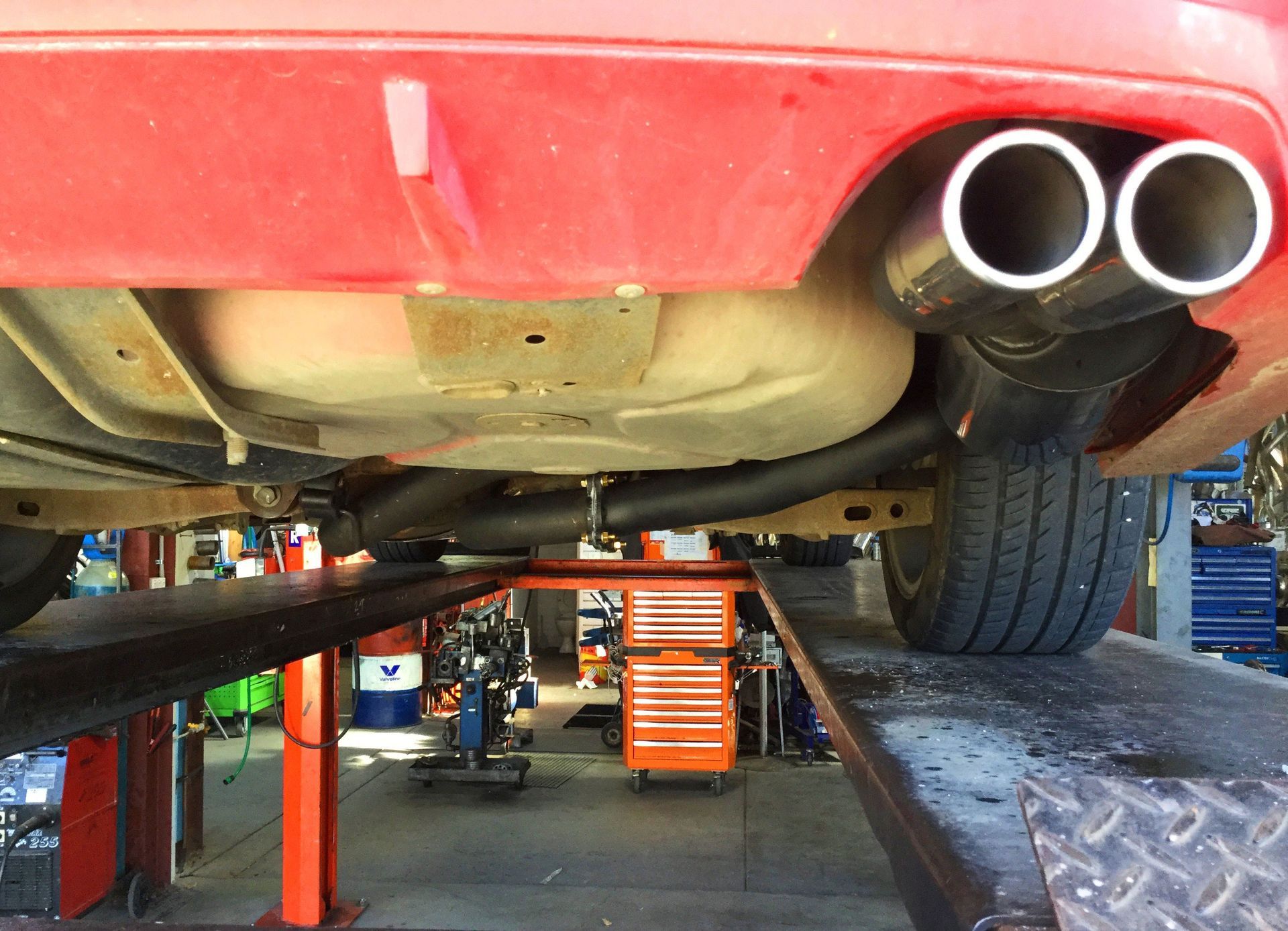 Red car's undercarriage with dual exhaust pipes raised on a lift in a shop.