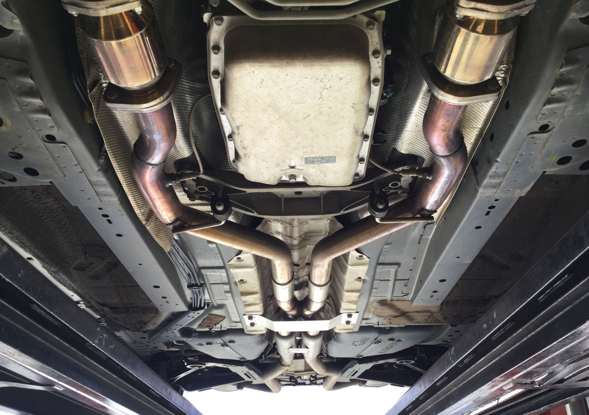 Undercarriage of a vehicle, showing exhaust system with metallic pipes, heat shields, and vehicle frame on a lift.