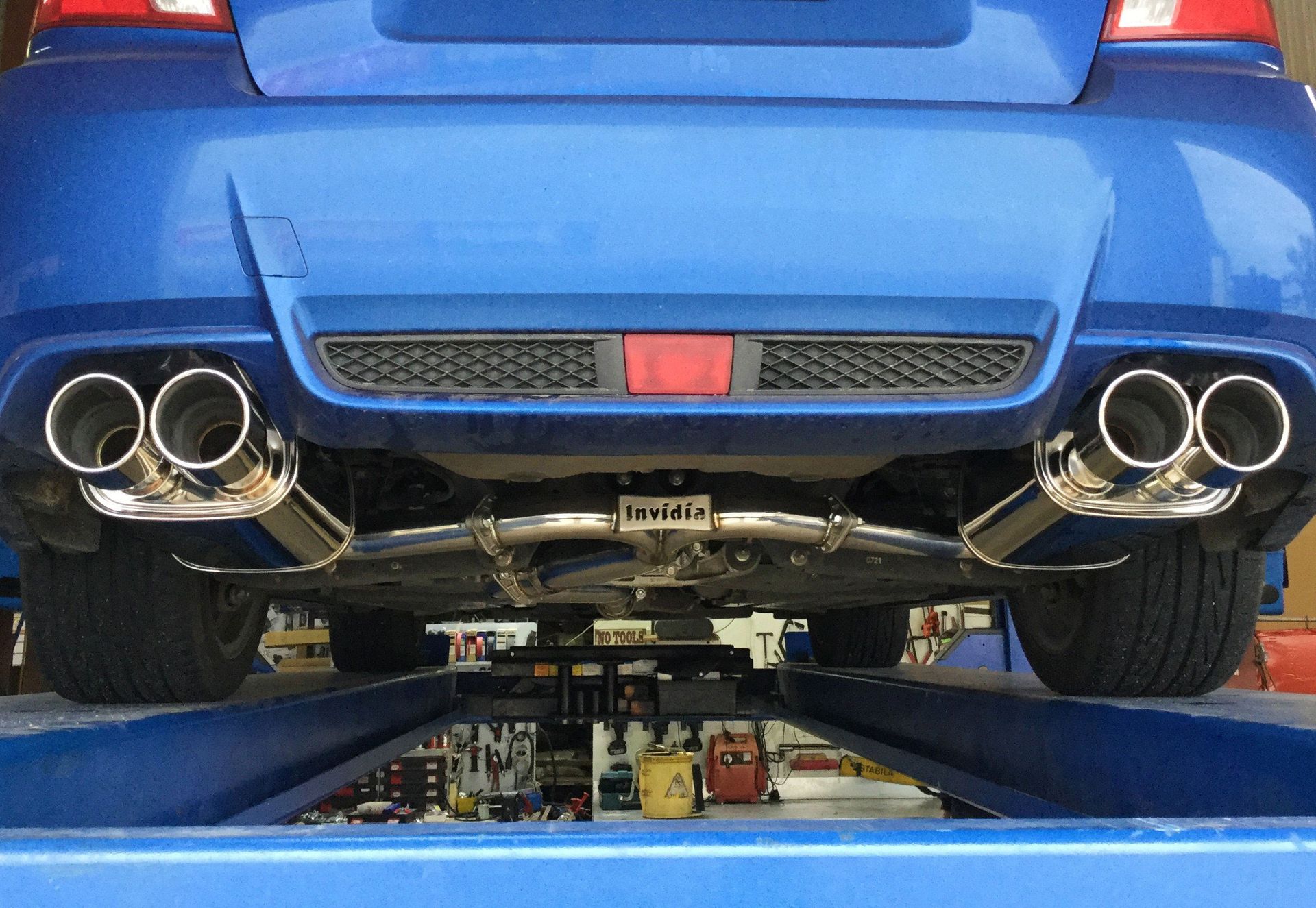 Blue car's undercarriage. Dual exhaust pipes and a red reflector are visible from below while the car is on a lift.