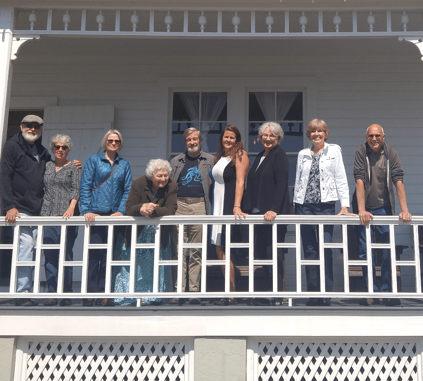 Group of people smiling, posing on a porch. White house backdrop. Sunny day.