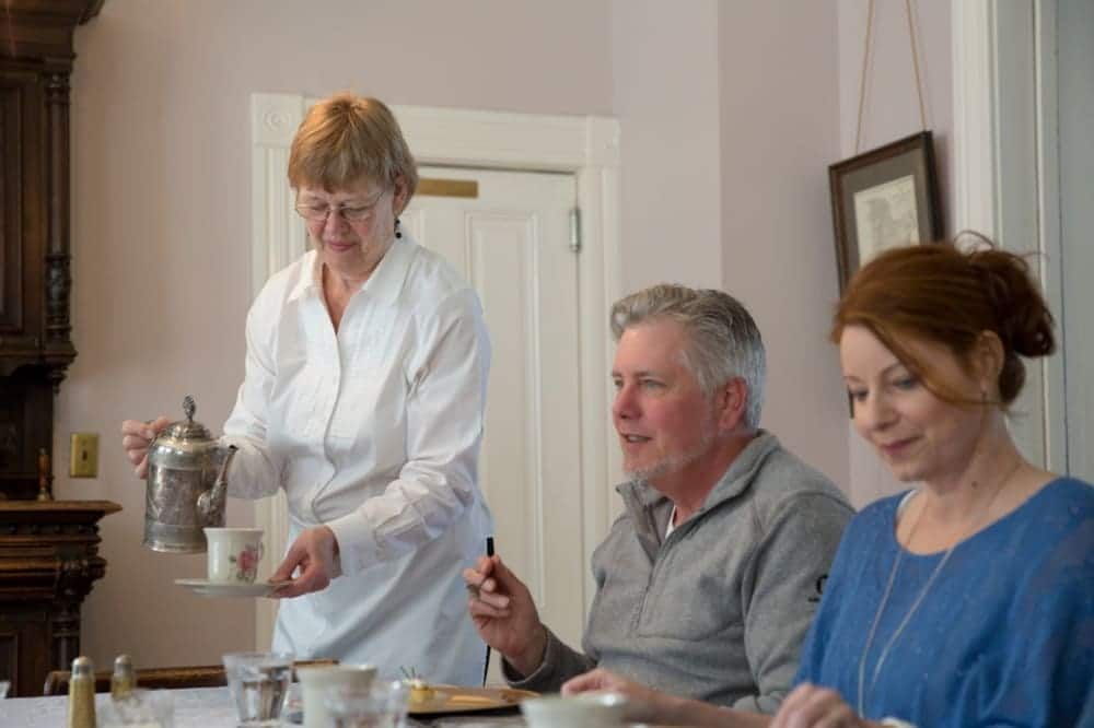 Woman pours tea for a couple seated at a table. Indoor setting with antique furniture.