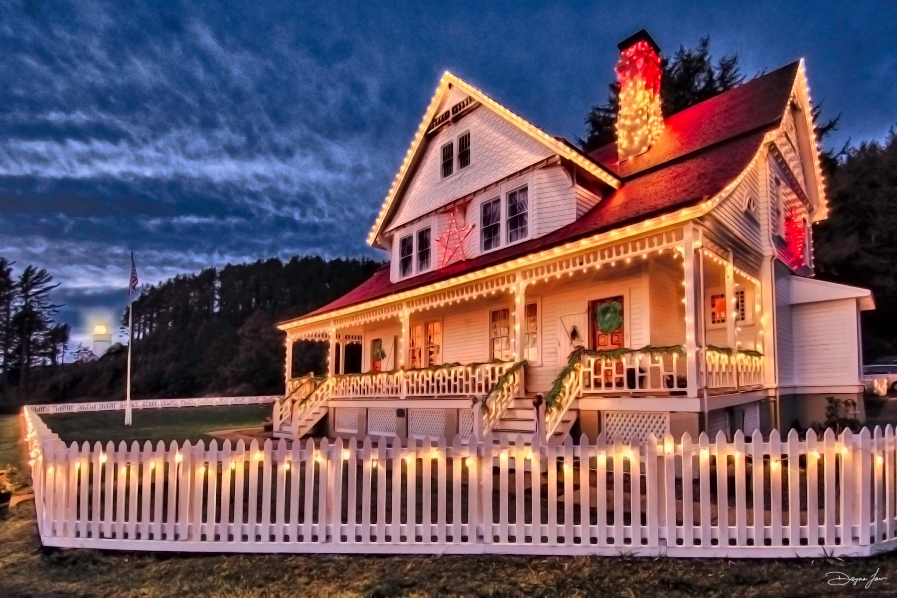 House with red roof, white fence, and lights. It's lit for Christmas in a night setting.