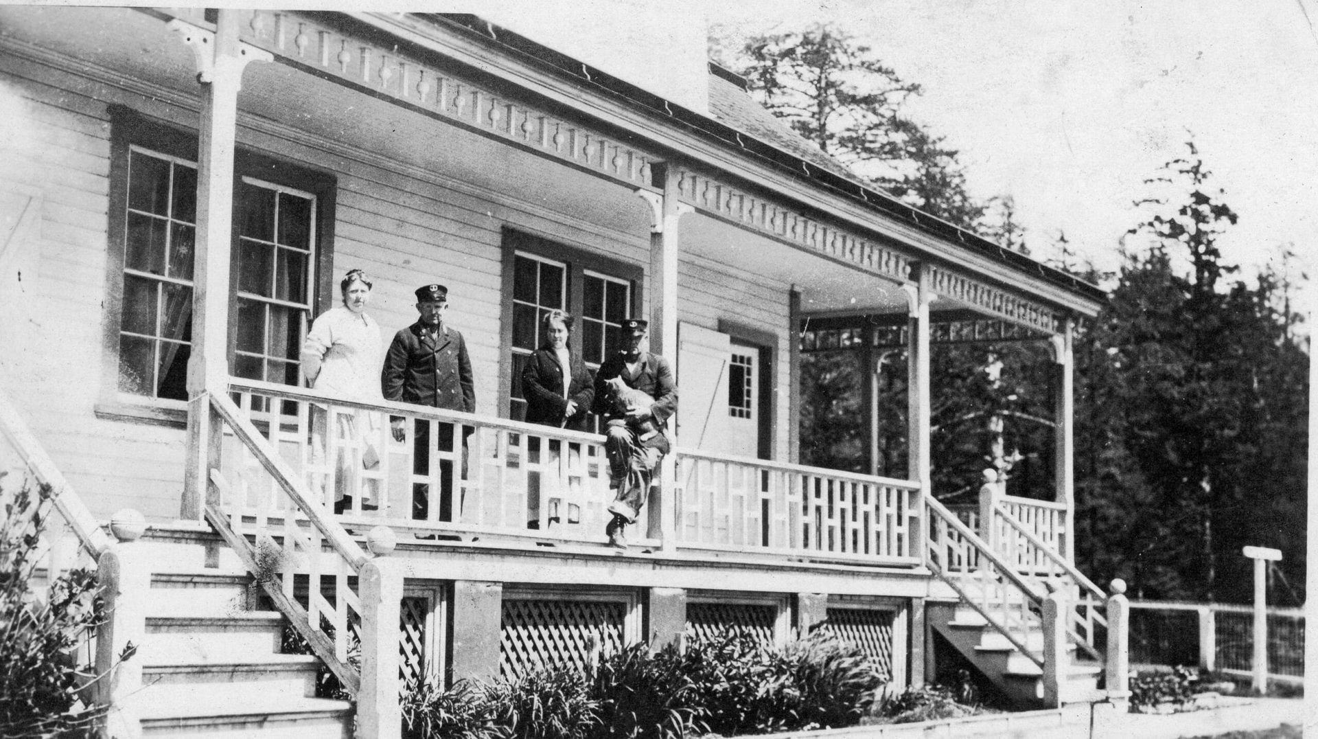 People on a porch of a white, wooden house with decorative trim.