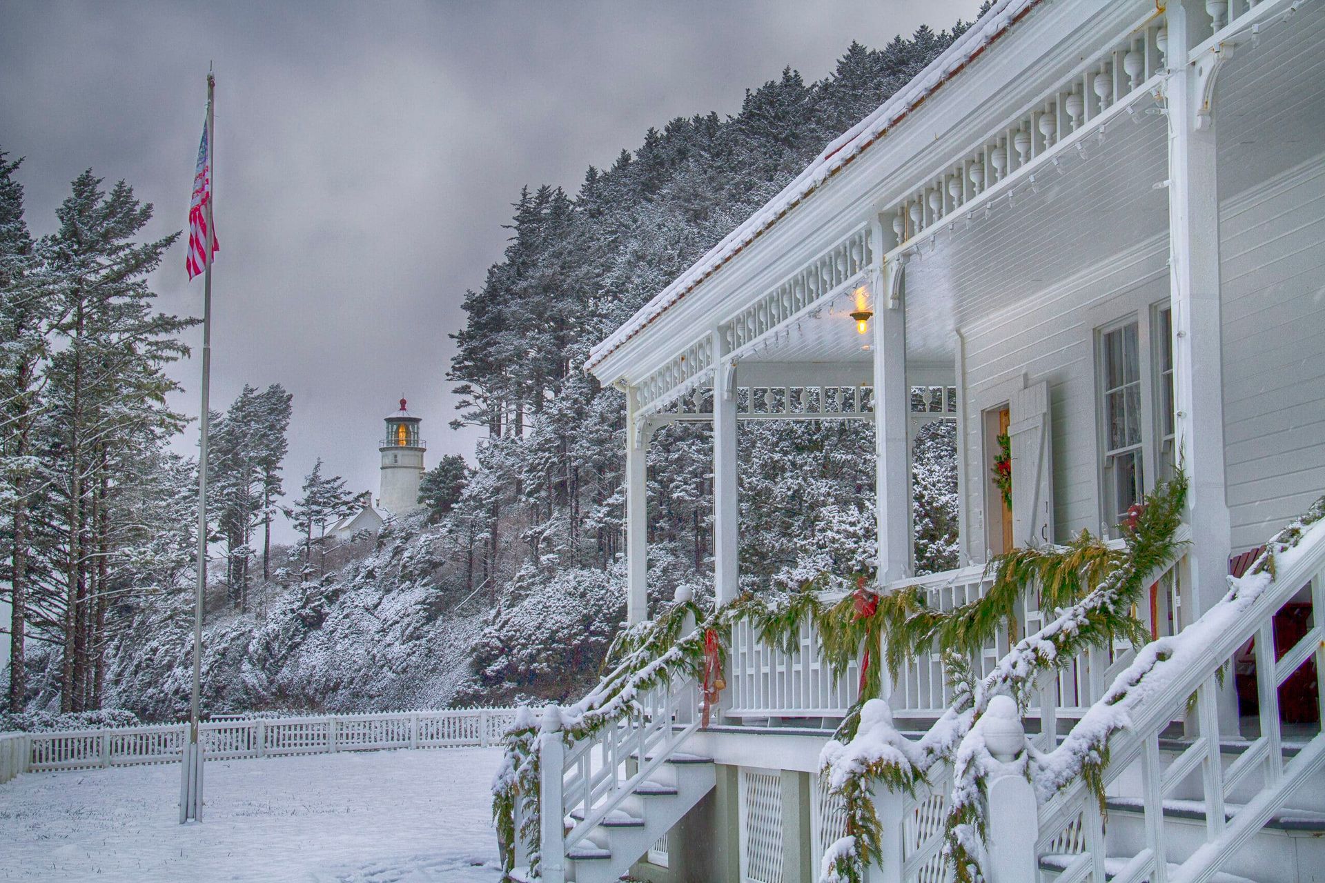 Snowy white house with porch and stairs, garland, flag, trees, and lighthouse in the background.