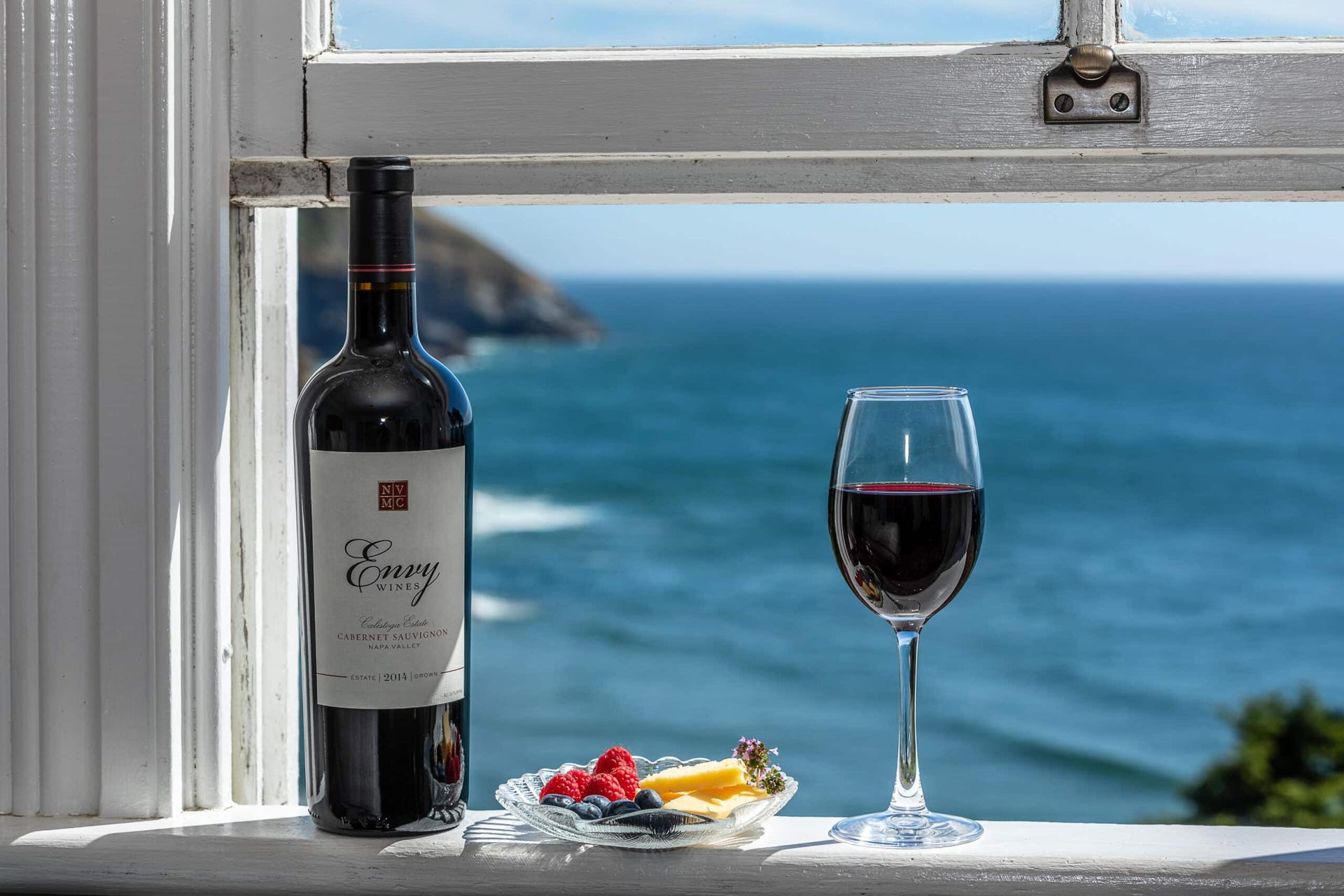 Bottle and glass of red wine, with fruit, on windowsill overlooking the ocean.