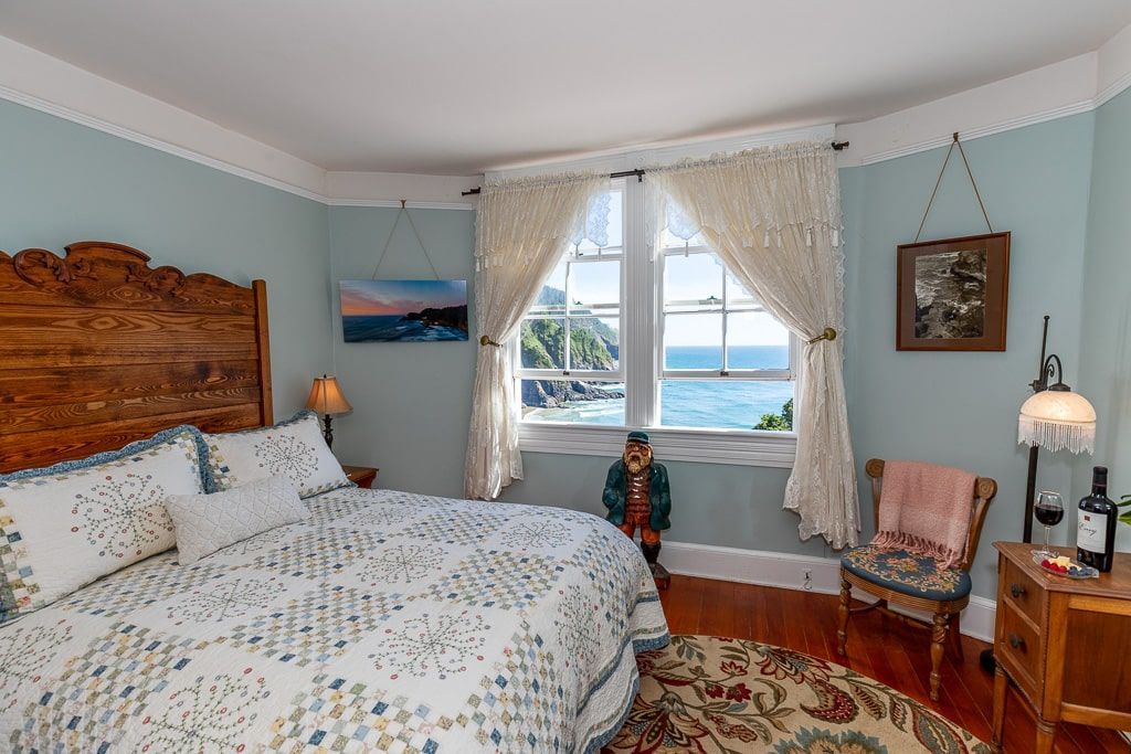 Bedroom with a bed, window overlooking ocean, small chair, and vintage furniture. Soft blue walls.