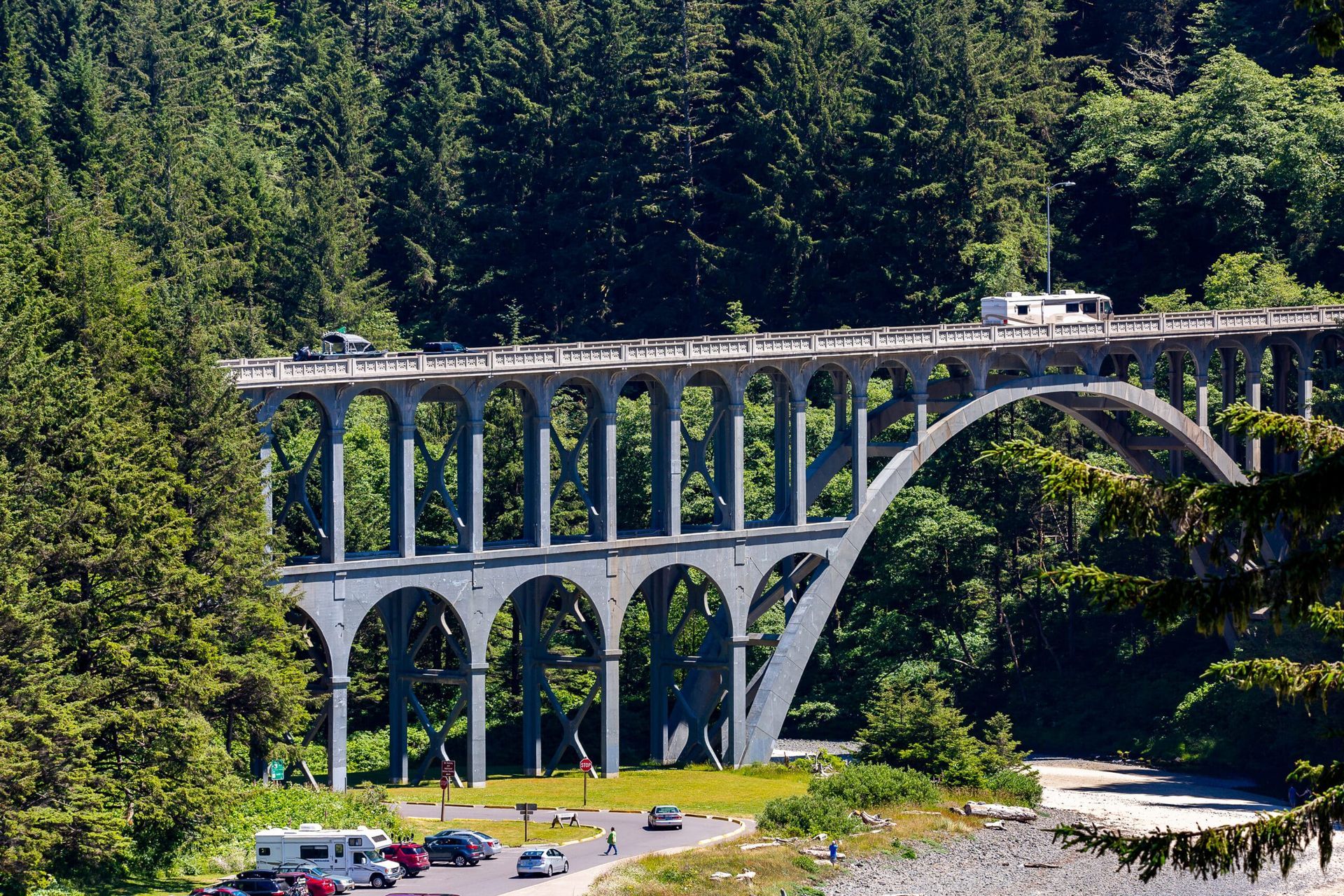 Scenic concrete arch bridge over a river, with cars and RVs parked nearby, surrounded by lush green trees.
