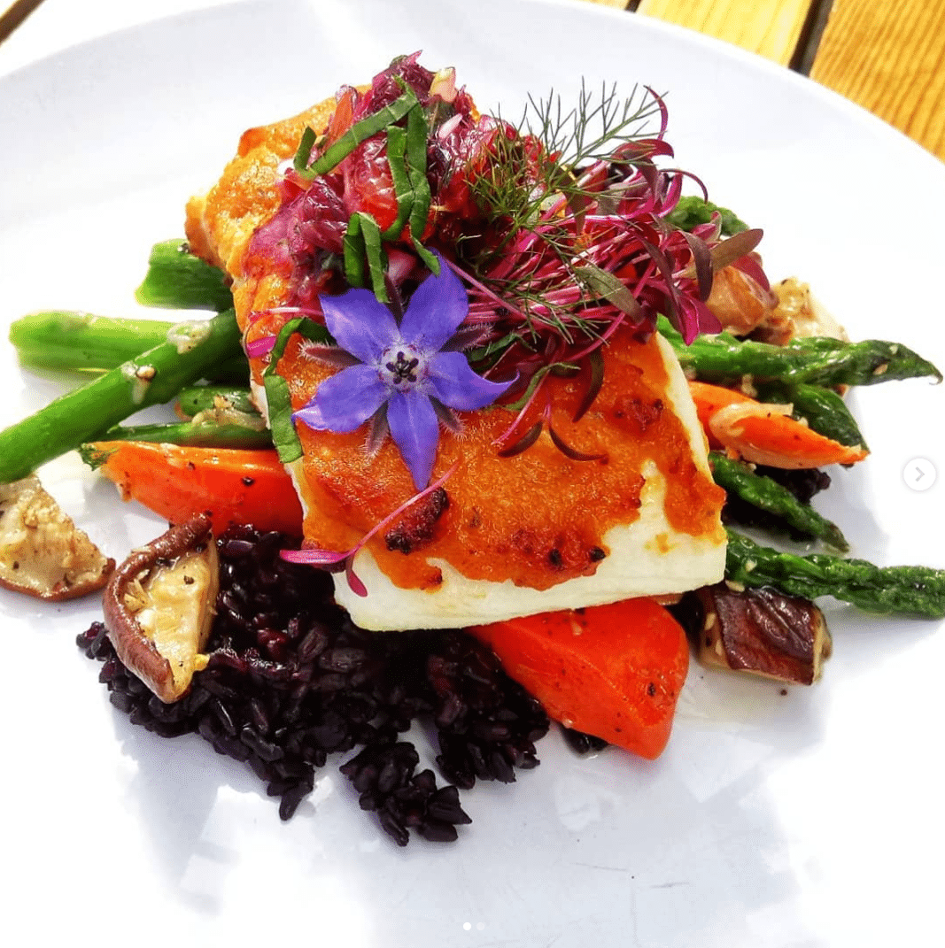 Pan-seared fish with purple flowers, asparagus, carrots, mushrooms, and black rice on a white plate.