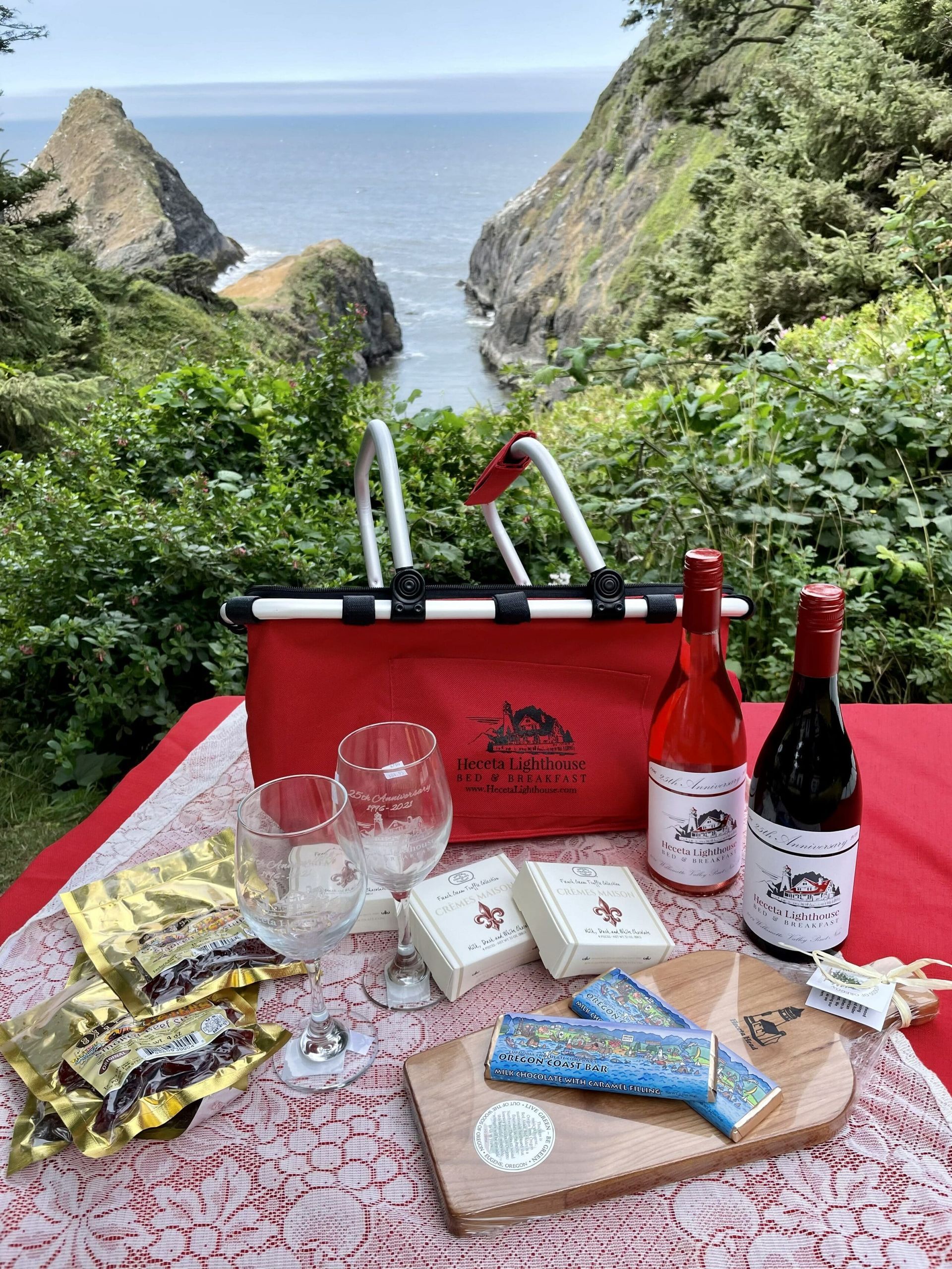 A picnic table with a bottle of wine , glasses , cheese , and a bag.