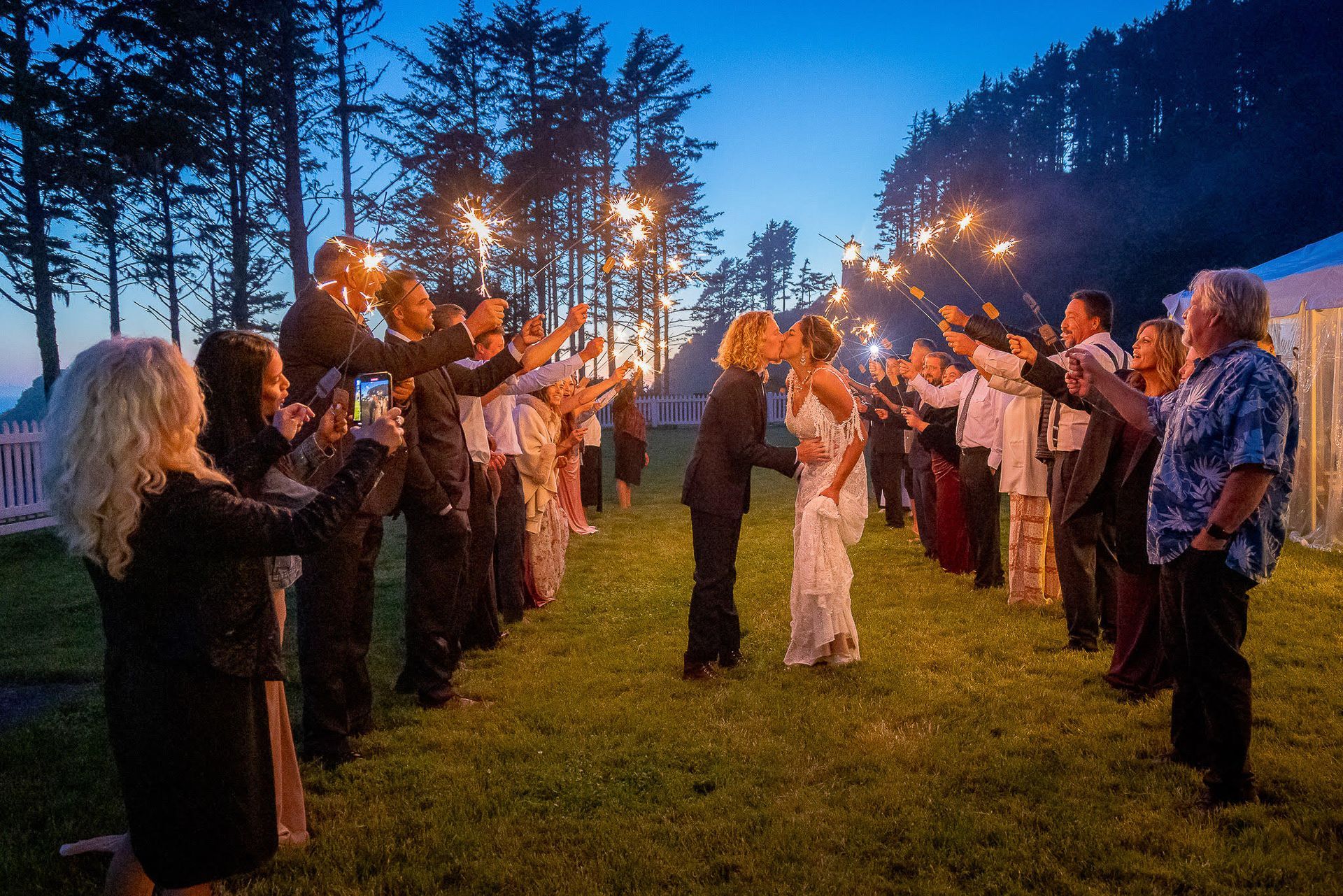 Newlyweds kiss while guests hold lit sparklers, creating a celebratory path. Evening outdoor wedding.