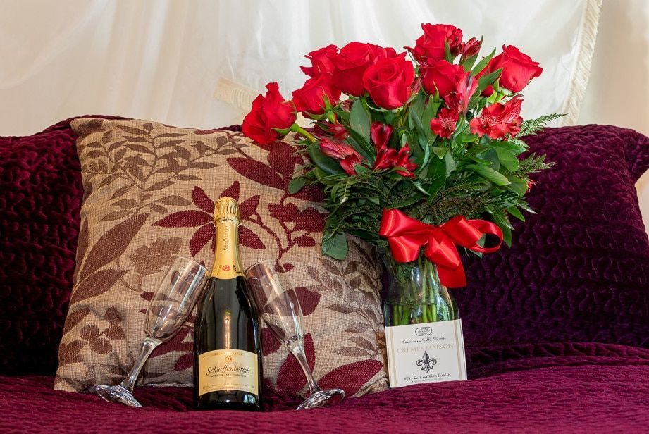 A bottle of champagne , a vase of red roses , and two glasses are on a bed.