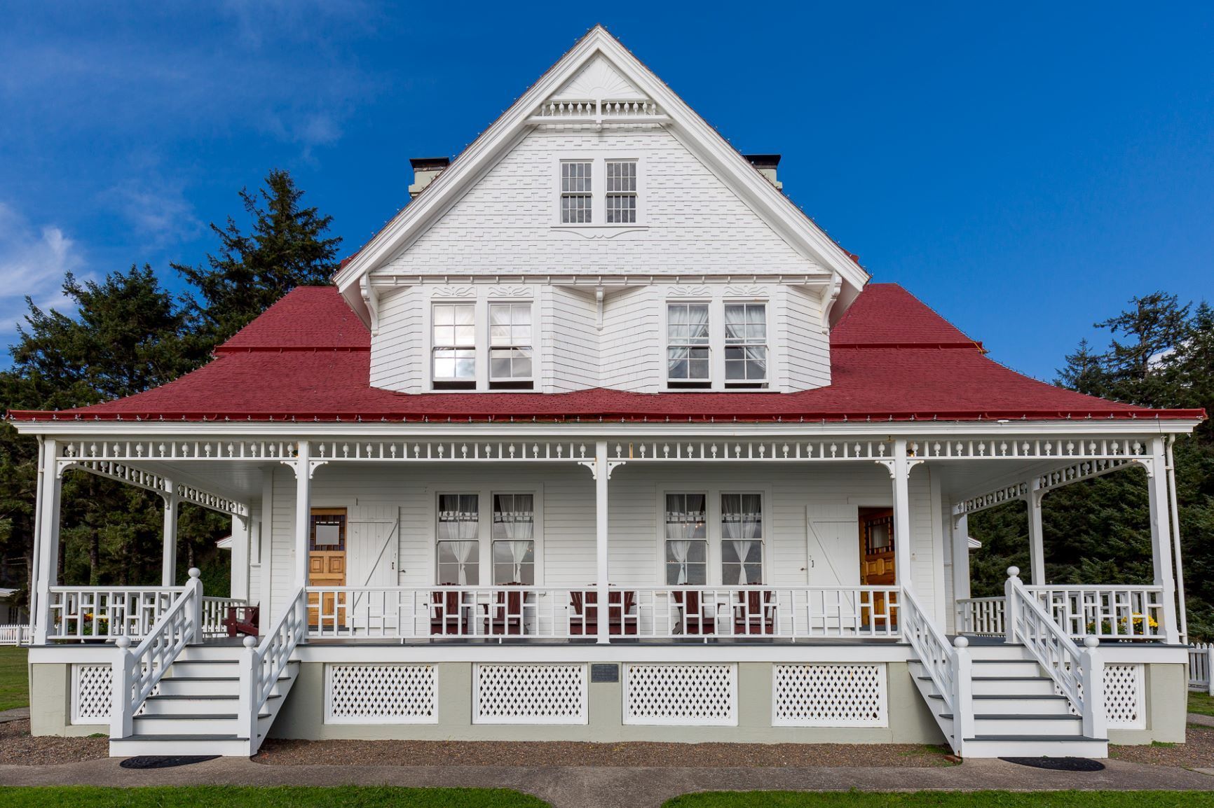 White two-story house with red roof and wraparound porch. Blue sky in background.
