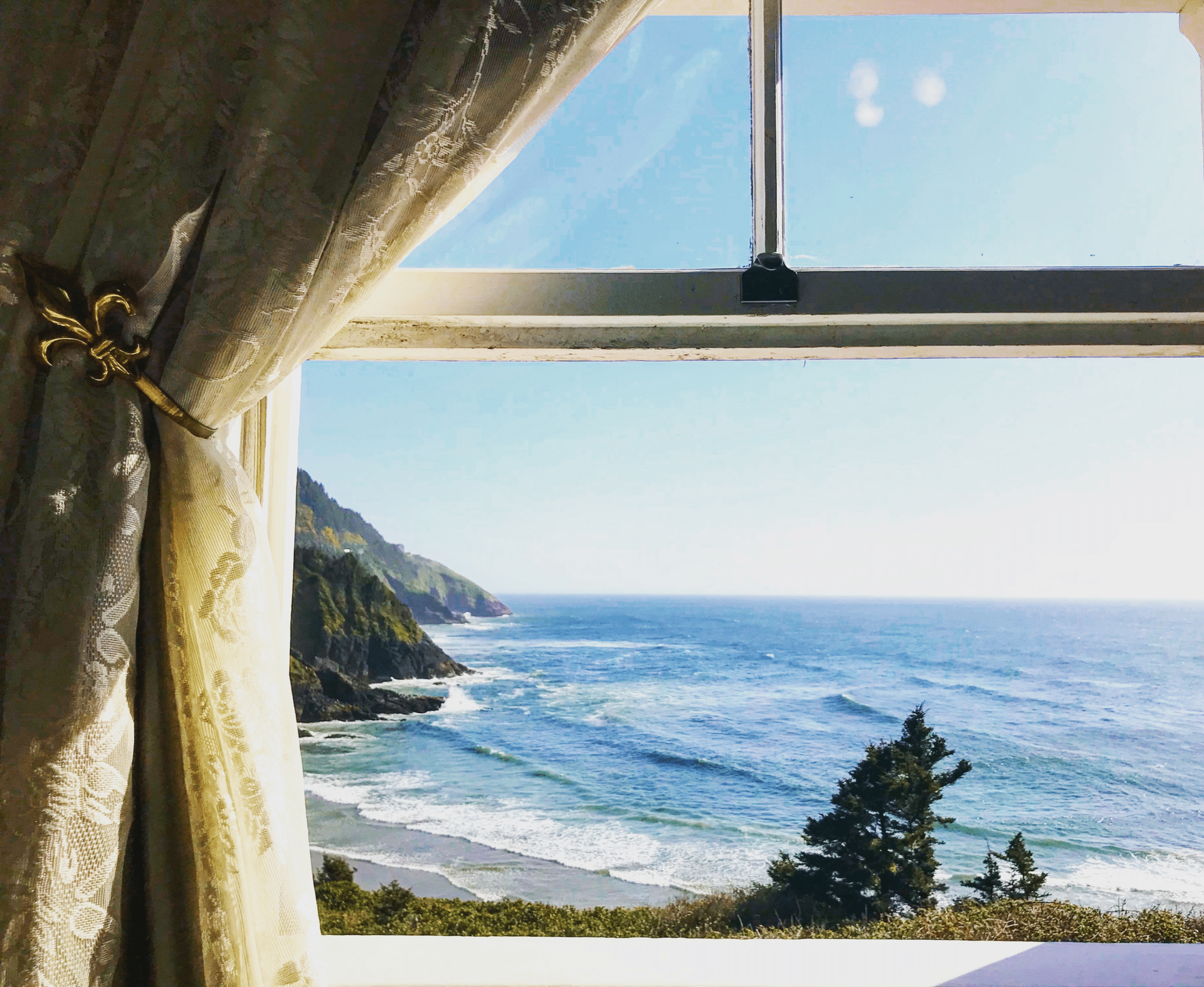 Window view of ocean and coastline on a sunny day; gold curtain tieback in the foreground.