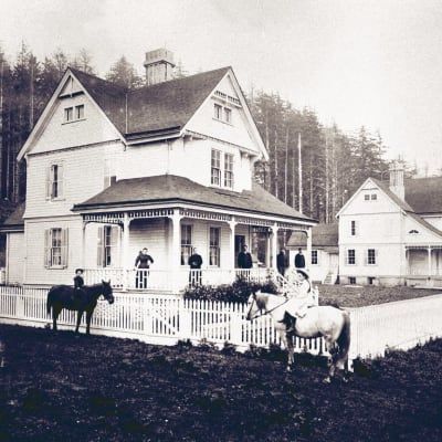 A black and white photo of people riding horses in front of a house