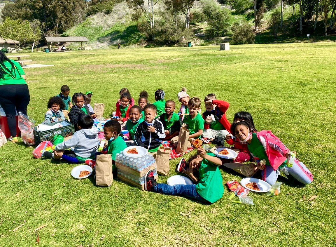 A group of young children in green shirts enjoy a picnic on a grassy hill on a sunny day.