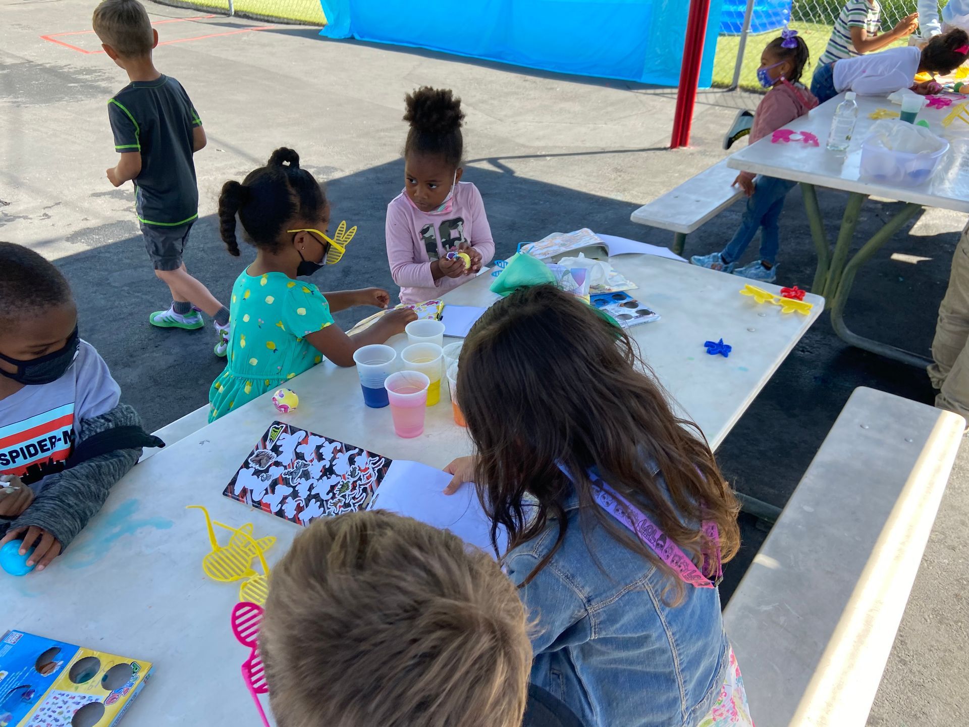 Children at an outdoor table doing a craft project, some wearing masks. They are using colorful liquids and small supplies.