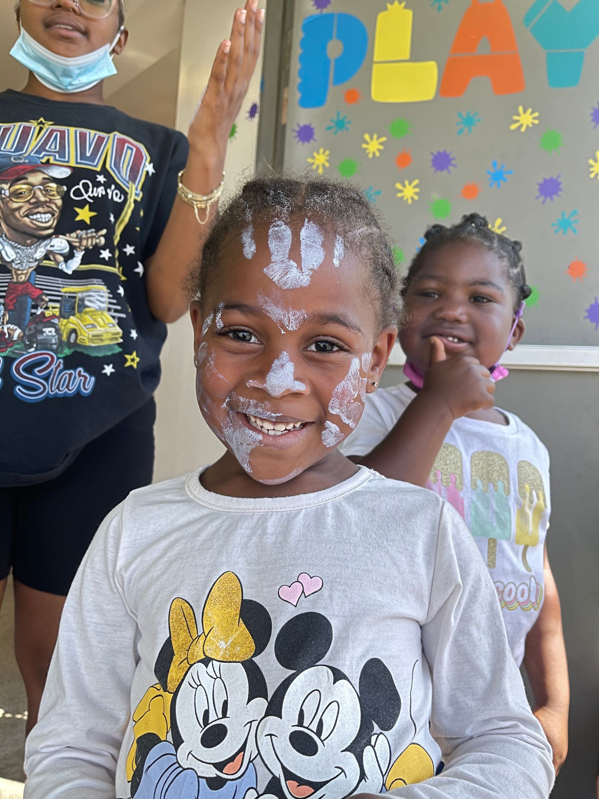 Girl with white paint on her face smiles, flanked by another girl and a woman with hand raised,