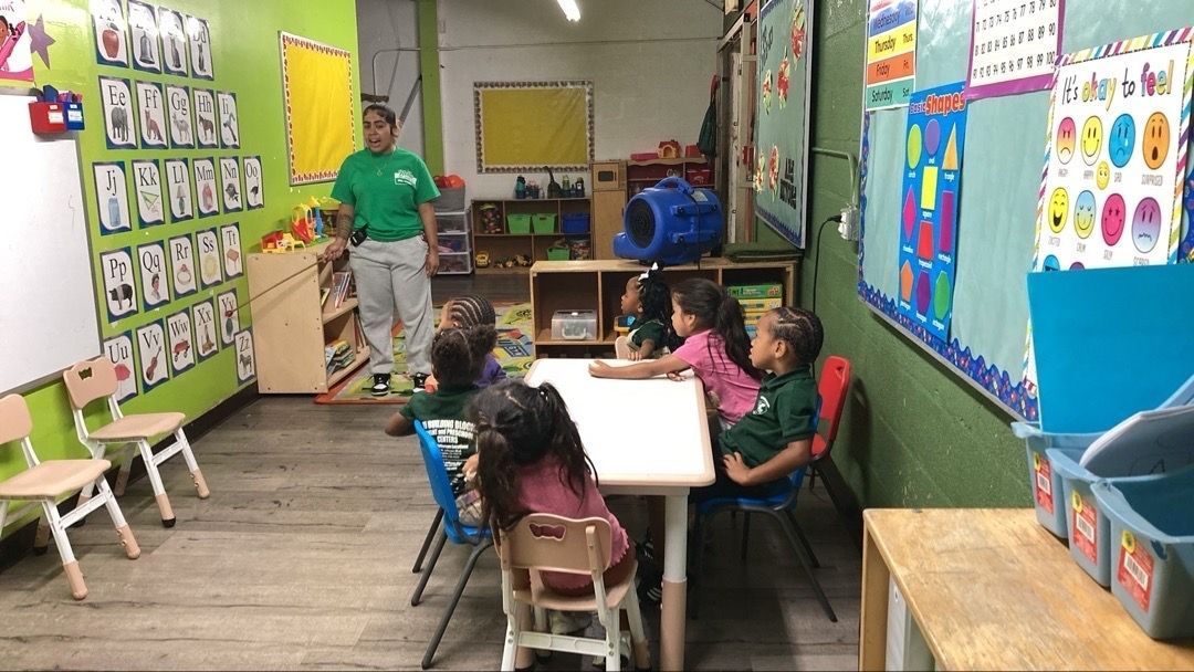 A teacher stands in a classroom with children sitting at a table; classroom features colorful decorations.