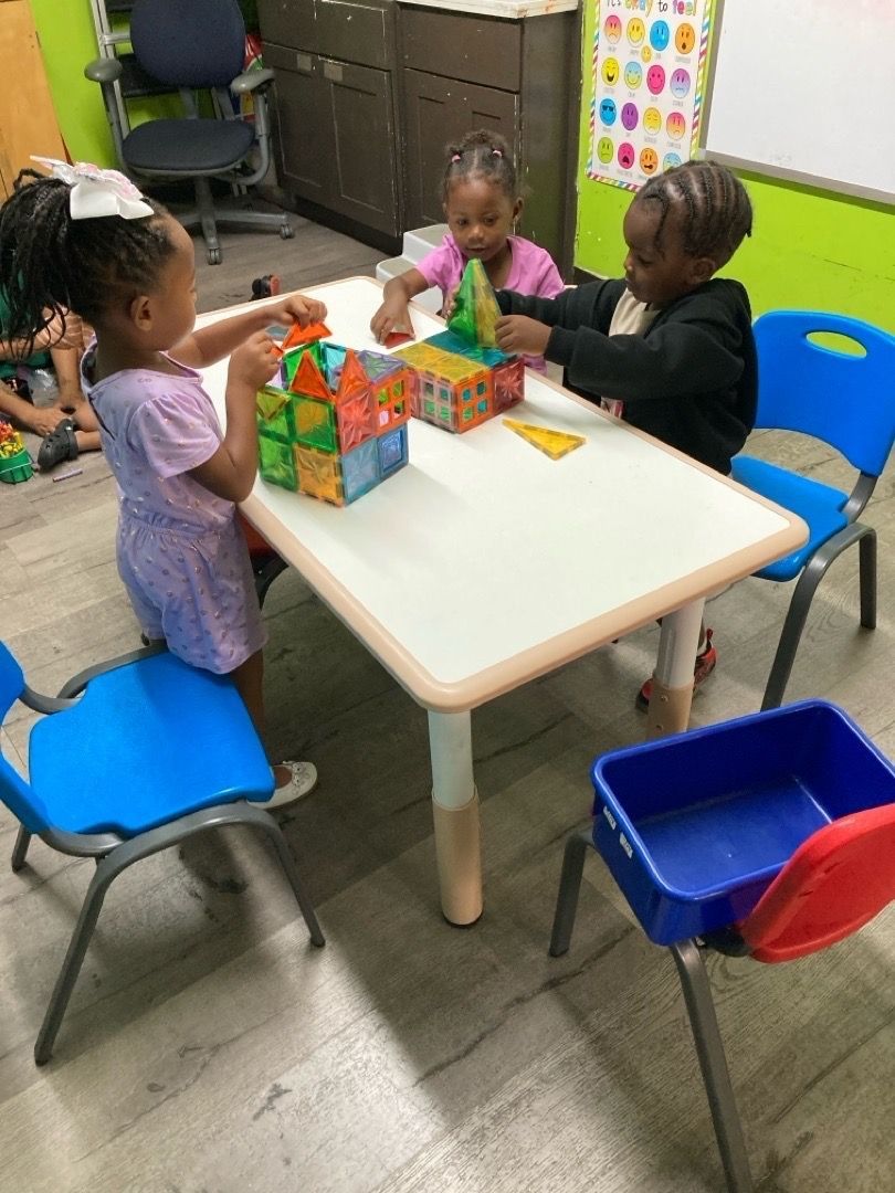 Three young children build structures with colorful magnetic blocks at a white table in a classroom.