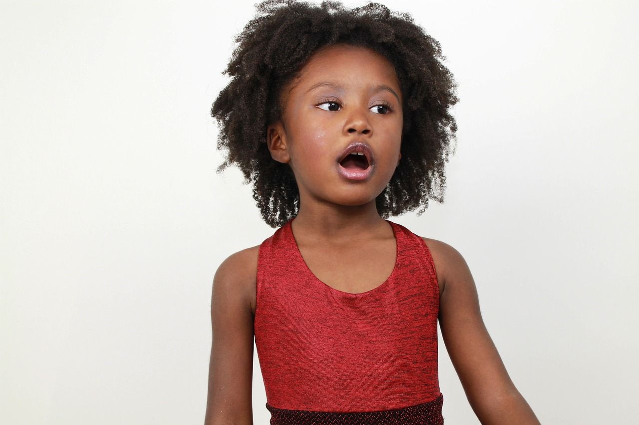 Young Black girl with curly hair, wearing a red sparkly top, looks surprised with mouth open on a white background.