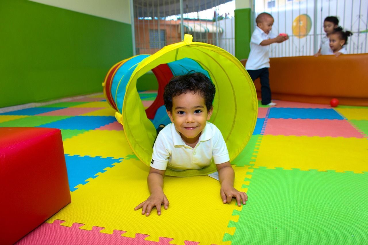 A smiling child crawls out of a colorful tunnel in a brightly colored play area.