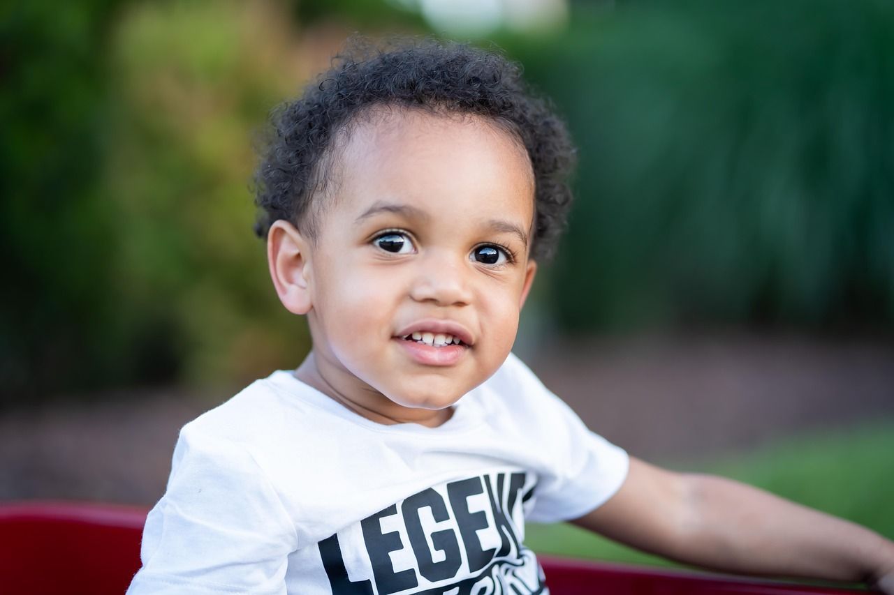 Young Black child with curly hair in a white t-shirt smiling, seated in a red wagon outdoors.