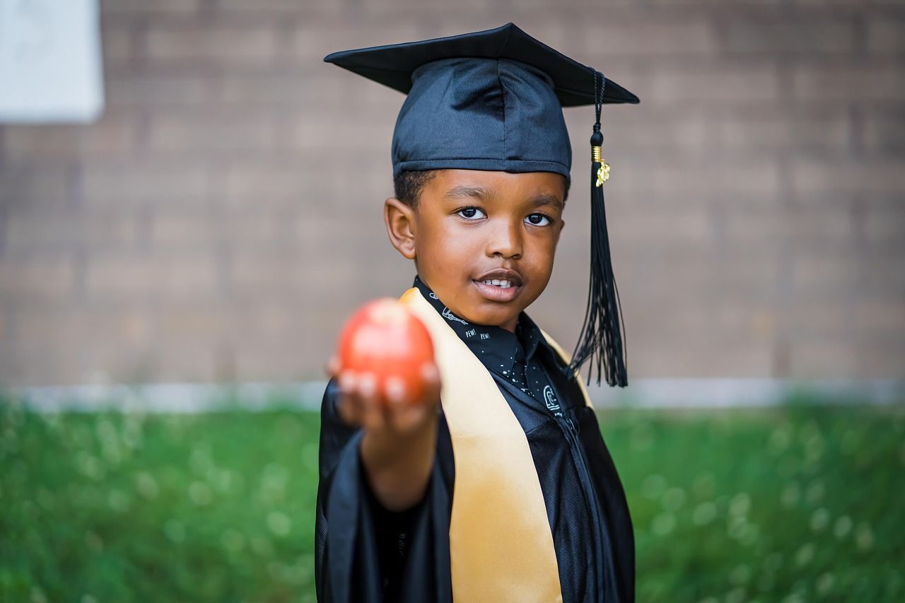 A young Black boy in a graduation cap and gown, holding out a red apple. Outdoors on a grassy lawn.
