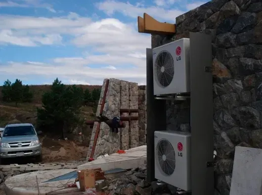 Unidades de aire acondicionado exteriores montadas en un muro de piedra junto a un camino rural y un coche aparcado.