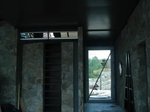 Pasillo estrecho sin terminar, con paredes de piedra vista y una luminosa puerta que da a una zona verde exterior.