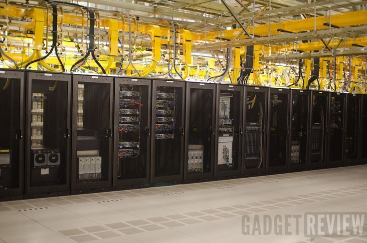 Rows of black server cabinets in a data center, with yellow overhead pipes.