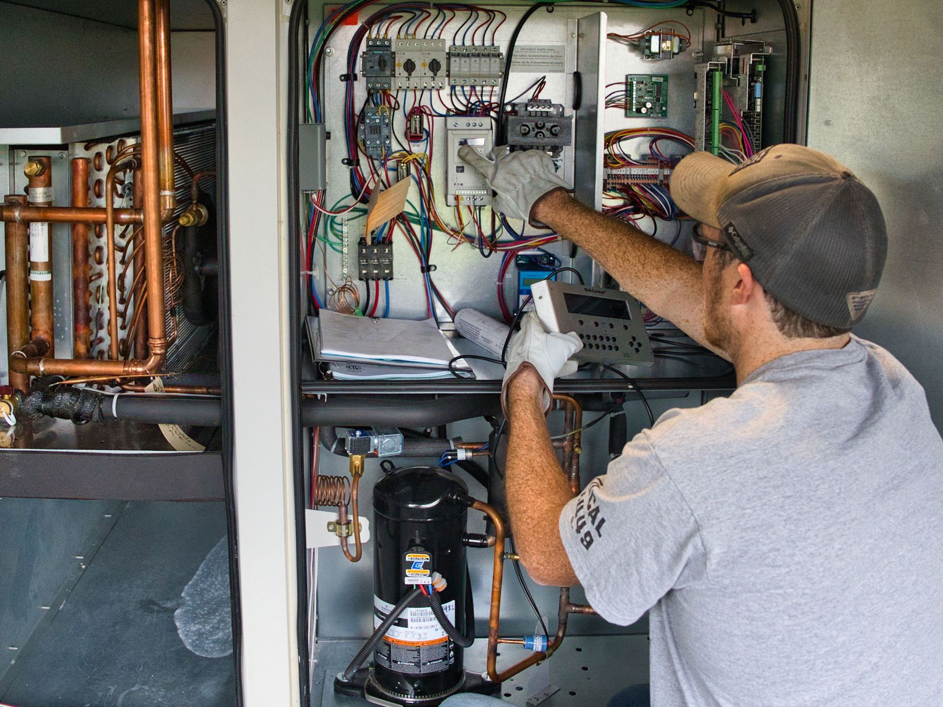 HVAC technician working on a unit, wearing gloves and a cap, inside a metal enclosure.