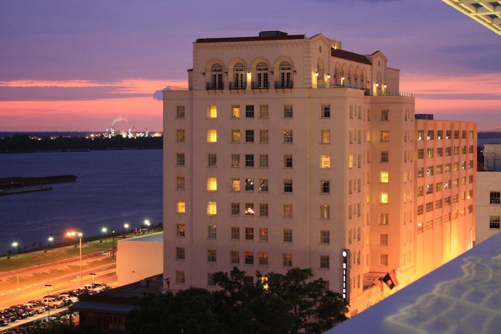 Tall, white hotel at dusk overlooking a river. Lit windows glow against the colorful sky.